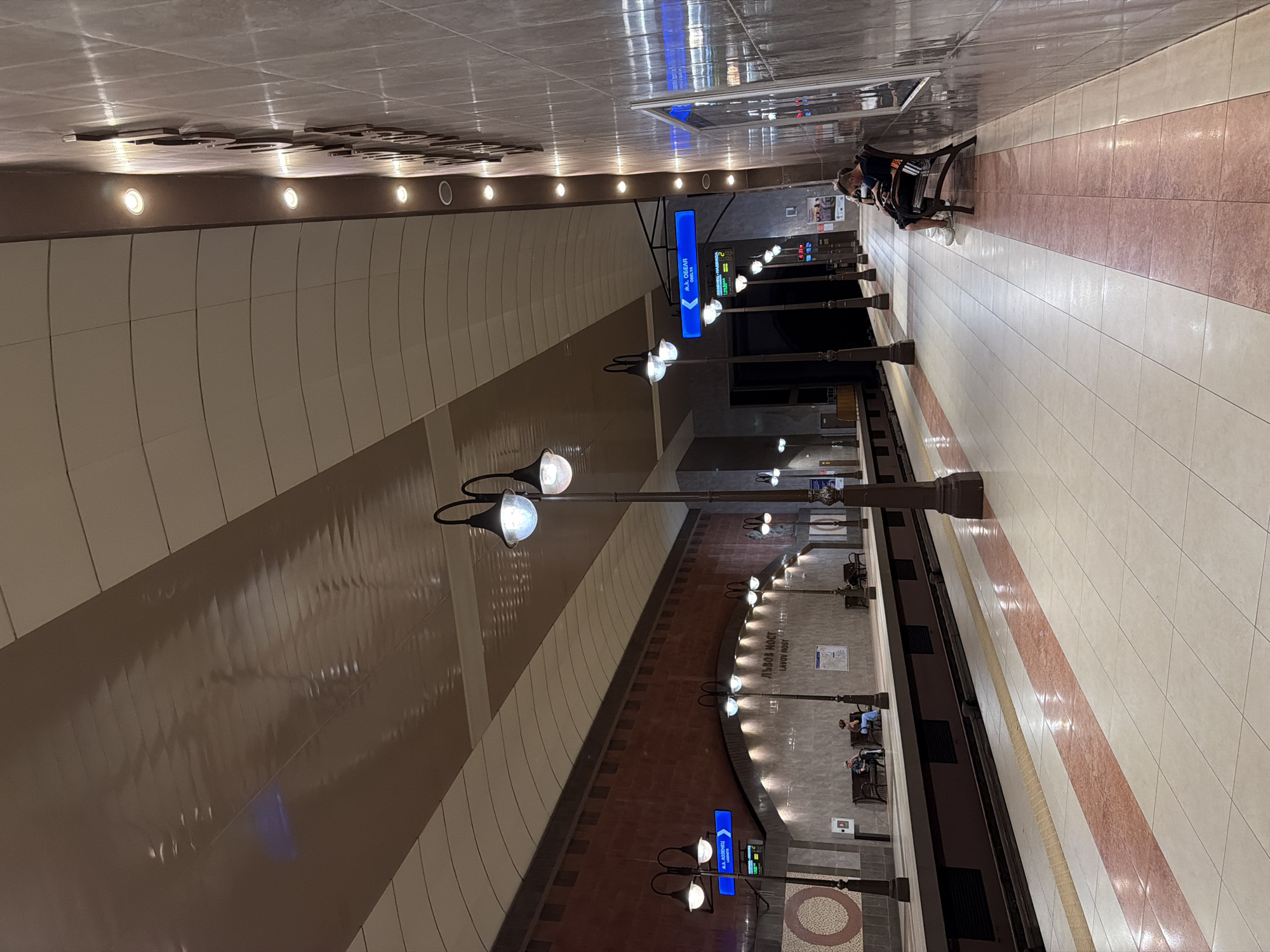 The image depicts a subway station platform in Sofia, Bulgaria. The platform is well-lit with overhead lighting and streetlamps. The walls are tiled with a marble-like pattern, and the ceiling is made of curved panels. There are benches along the platform for passengers to sit on. In the distance, a digital sign displays station information. Several people are visible on the platform, waiting for trains or resting on the benches. One person is seated on a bench, looking down at their phone. Another person is standing near the sign, possibly checking the train schedule. The platform floor is tiled with a pattern of white and red tiles.