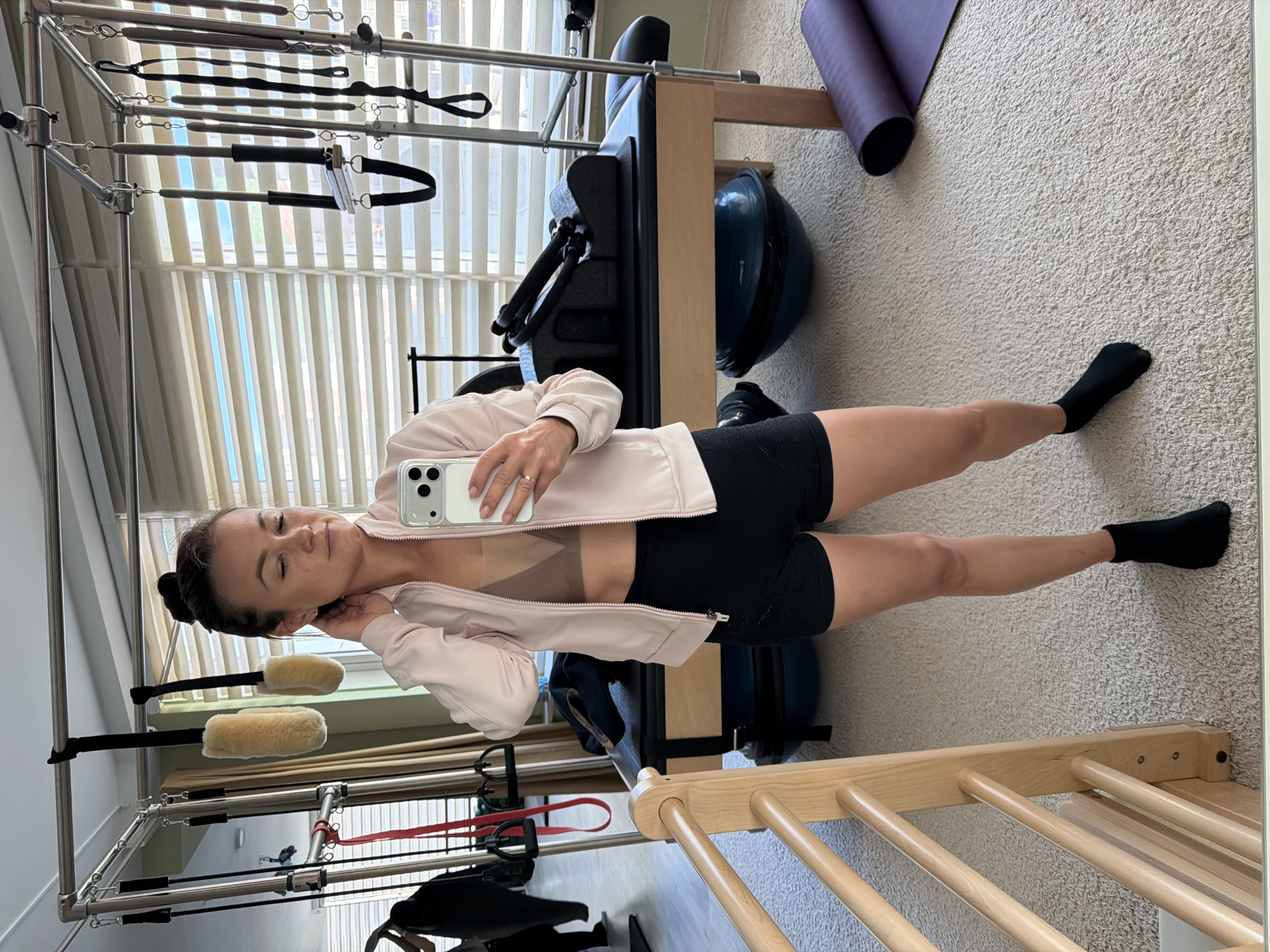 A woman is standing in a room that appears to be a fitness studio or home gym. She is wearing a light pink jacket, tan athletic bra, and black athletic shorts. She is wearing black socks. She is holding a smartphone in her right hand. The smartphone appears to be an iPhone.
Behind her is a pilates reformer machine. Next to the reformer machine, there is a balance ball. Further in the background, a pilates mat is rolled up, leaning against the wall.
A window is visible in the background. The window blinds are closed. The location is within a city, possibly Budva, Montenegro. The environment is well lit.