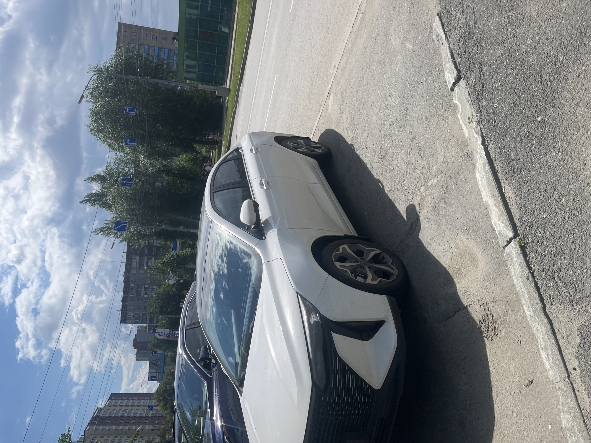 A white sedan is parked on a cracked concrete curb on the side of a paved road. The car's front is facing right, and its grille has a distinctive design with horizontal lines. Another dark-colored vehicle is parked to the left of the white sedan. In the background, there are multi-story residential buildings, trees, and a bright blue sky with white clouds. Several blue traffic signs indicating direction are visible among the trees. A green structure with glass panels is located further down the road on the right. The scene appears to be in an urban environment, possibly Chelyabinsk, Russia, given the architectural style of the buildings and the presence of street signs that are consistent with Russian road signage. The lighting suggests it is daytime.