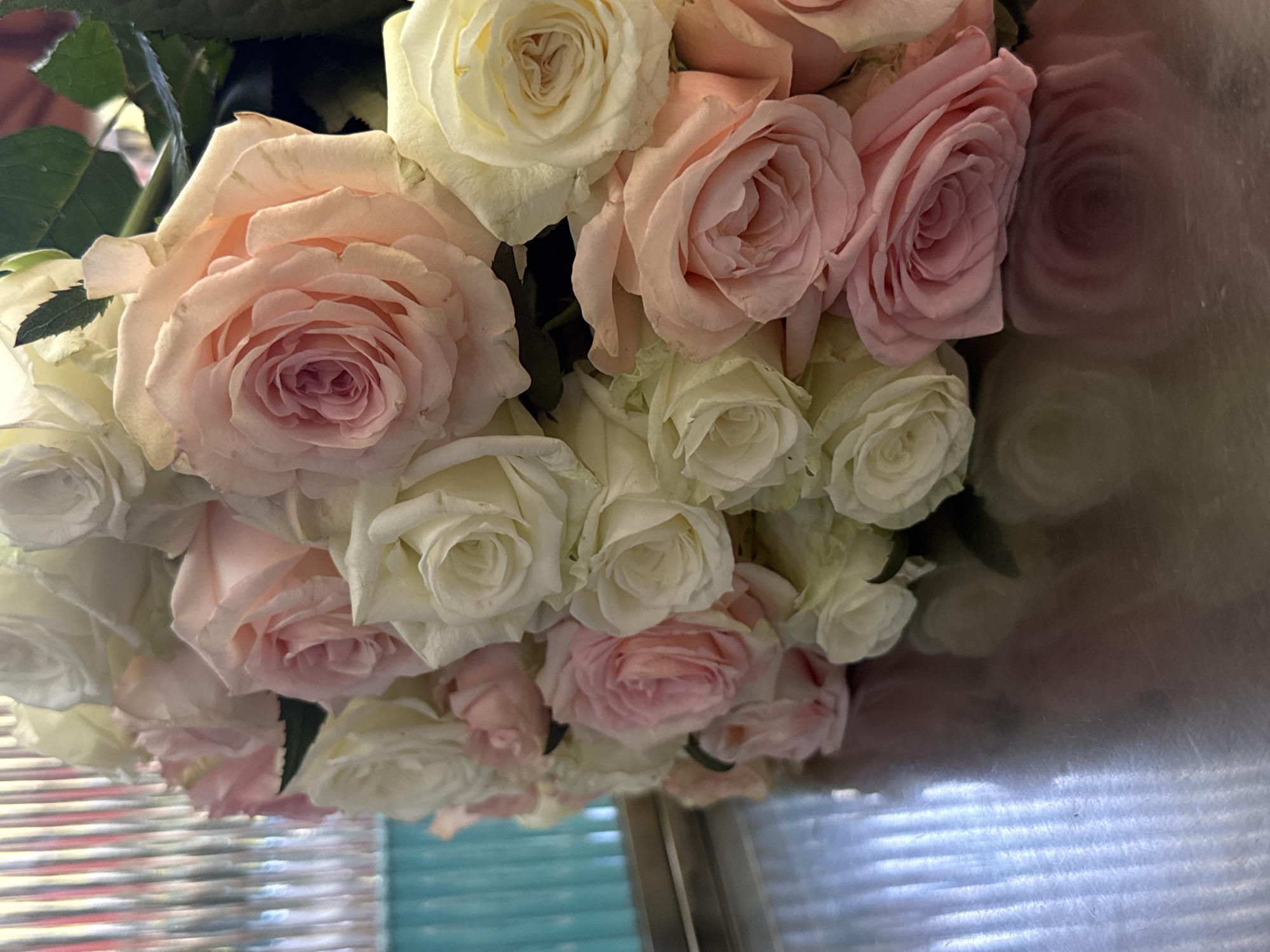 A bouquet of mixed pink and white roses is arranged on a reflective surface, possibly a counter or tabletop. The bouquet comprises numerous roses, densely packed together, with varying shades of pink and white. The reflection indicates the setting may be indoors, near a window with blinds. There is no indication that the image was taken in Yerevan, Armenia.