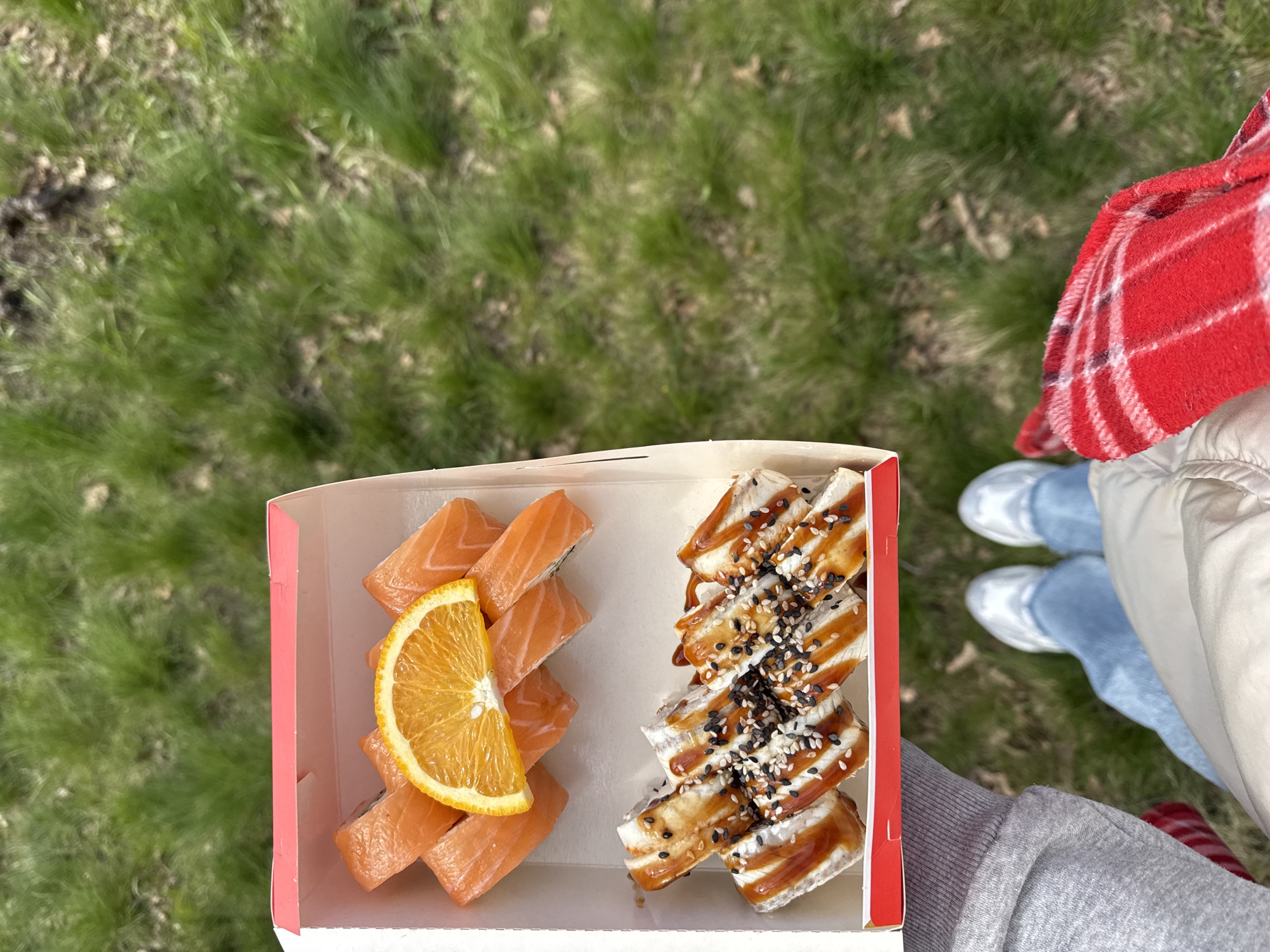 A person stands outdoors on grass, holding a takeout container of sushi. The container is divided into two sections: one contains salmon sushi rolls with a slice of orange on top, and the other contains sushi rolls topped with eel and drizzled with a sauce and sprinkled with sesame seeds. The person is wearing jeans, white sneakers, and a grey sweatshirt with a white jacket visible, and a red and white scarf is draped over their shoulder. The background is a blurred view of green grass. The location is an unknown area within the city of Unknown, Unknown.