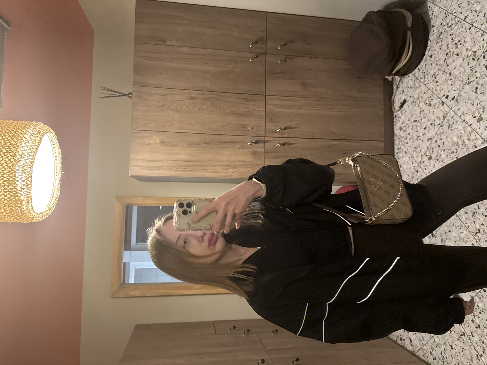 The image shows a young woman taking a selfie in what appears to be a locker room or dressing area. She is wearing a black jacket with white stripes on the sleeves and black pants. She is holding an iPhone in her right hand, which is decorated with flowers. The phone is partially covering her face. She is fair-skinned with blonde hair and is wearing a necklace. A Guess handbag with a chain strap hangs from her left shoulder.
The background includes wooden lockers and cabinets with metal handles. A brown hat sits on top of a cabinet in the upper right corner of the frame. The floor is tiled with a black and white pattern. In the upper left corner, there's a portion of a wall painted a light pink hue, and a wicker lampshade is visible. The mirror reflects a window and what may be a white tiled wall, suggesting the presence of a shower or bathroom area beyond the locker room. Rostov-on-Don, Russia is not identifiable in this image.