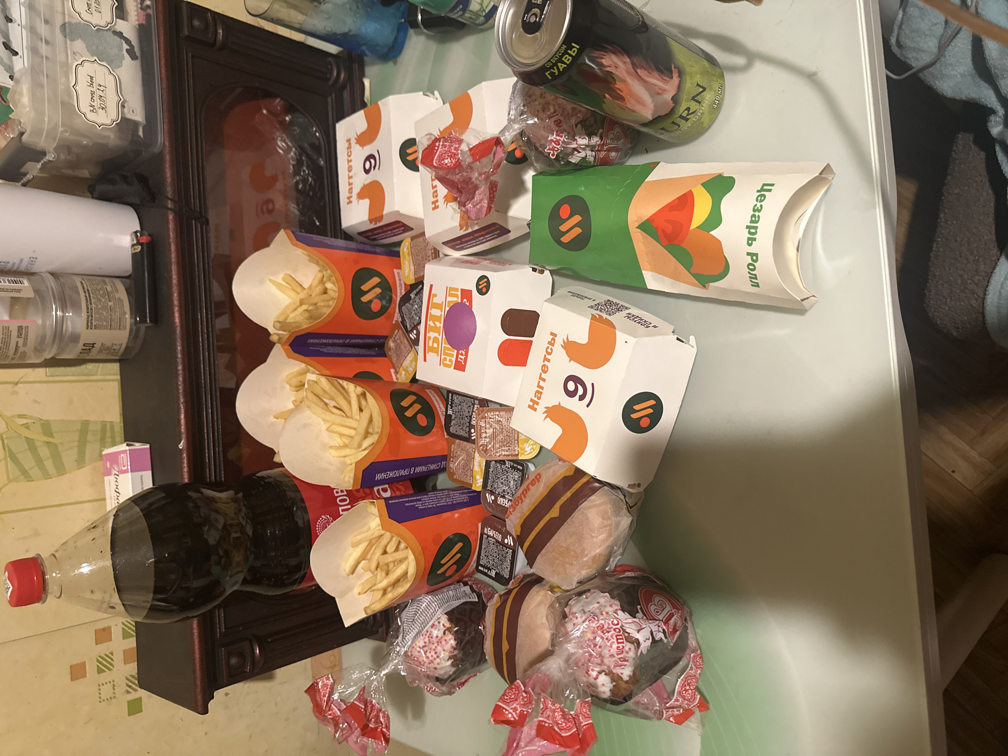 The image shows a table covered in fast food items. There are several boxes of "Naggets" (nuggets), containers of french fries, hamburgers, a "Caesar Roll," a bottle of Coca-Cola, a "Big Daddy" candy bar, and a can of "BURN" energy drink with guava flavor. All text is in Russian, therefore, it is assumed that the location is in a Russian-speaking area, likely Moscow, Russia.