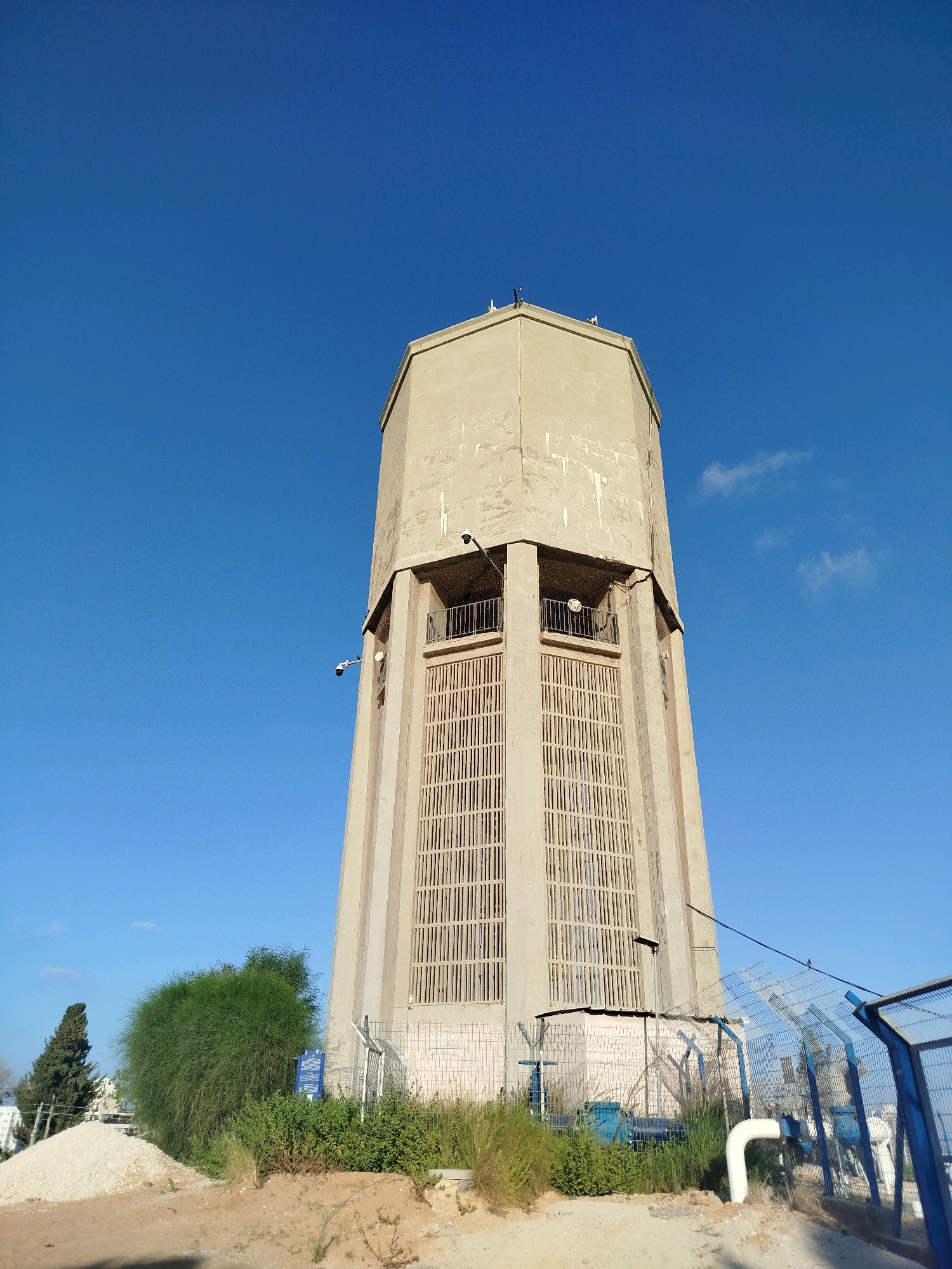 The image shows a tall, octagonal, concrete water tower located in Ashkelon, Israel. The tower is pale beige and shows signs of weathering. It has a decorative grated section around its upper half, with balconies visible behind the grating. There are security cameras mounted on the tower's exterior.
At the base of the tower, there is a blue metal fence surrounding industrial equipment, including large blue pipes and a white pipe. Vegetation, including green shrubs and dry grasses, grows around the base. To the left of the tower, there is a pile of light-colored gravel or sand, a tall, slender green tree, and a smaller, bushy green tree. A blue sign is visible on a post near the fence. The sky is a clear, bright blue with a few wispy white clouds. The scene appears to be a functional industrial site, possibly a water treatment or distribution facility.