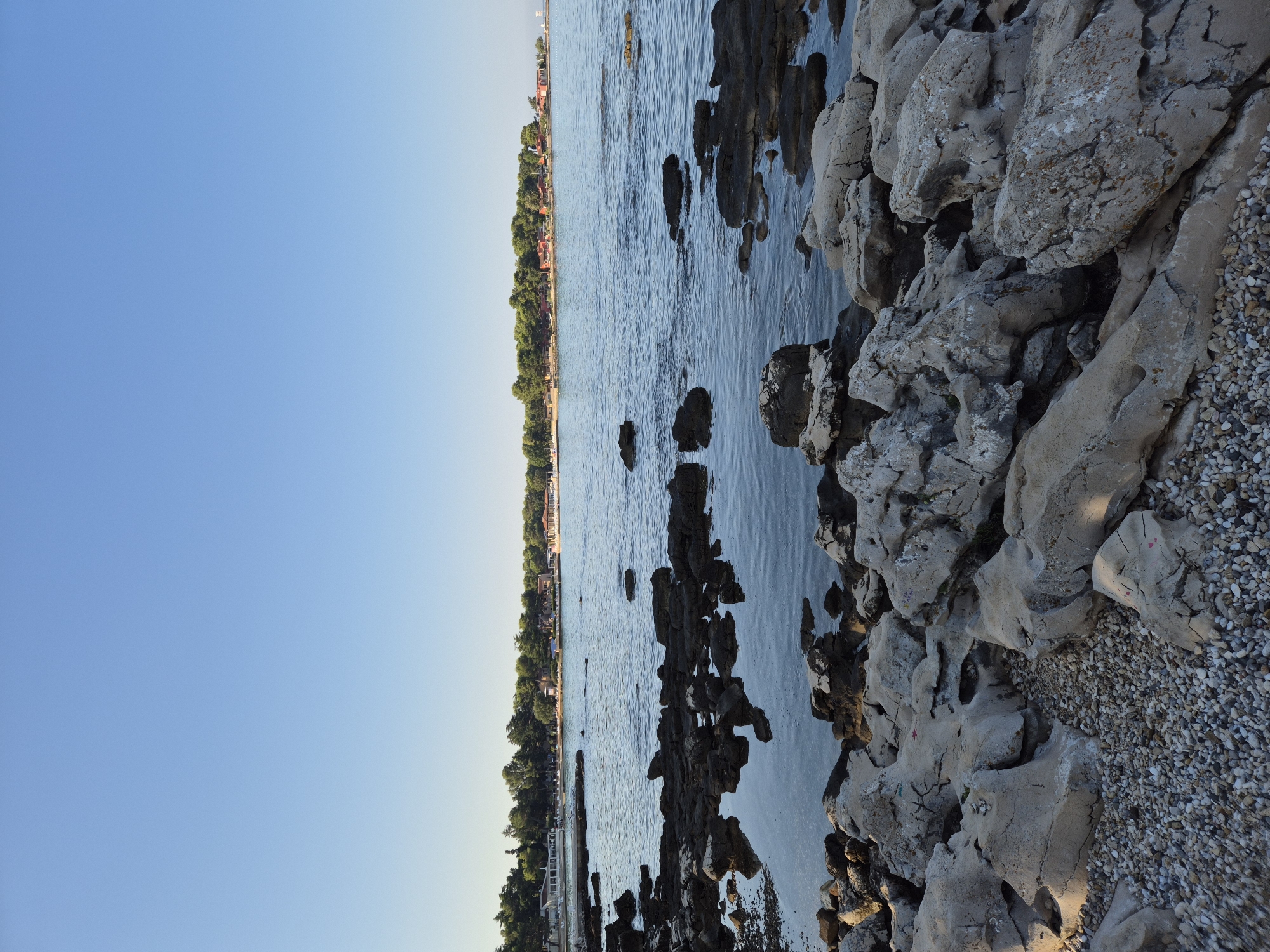 The image shows a rocky shoreline with the sea and a town in the background. The sky is clear and blue. The foreground is dominated by large, grey rocks and pebbles. In the middle ground, the calm sea laps against the rocks, with reflections visible. In the background, a line of green trees separates the water from a town with red-roofed buildings and some white structures. The scene appears to be a coastal area in Monterol, Croatia, likely a public beach or recreational area, captured from a low angle. There are no people visible in the frame.