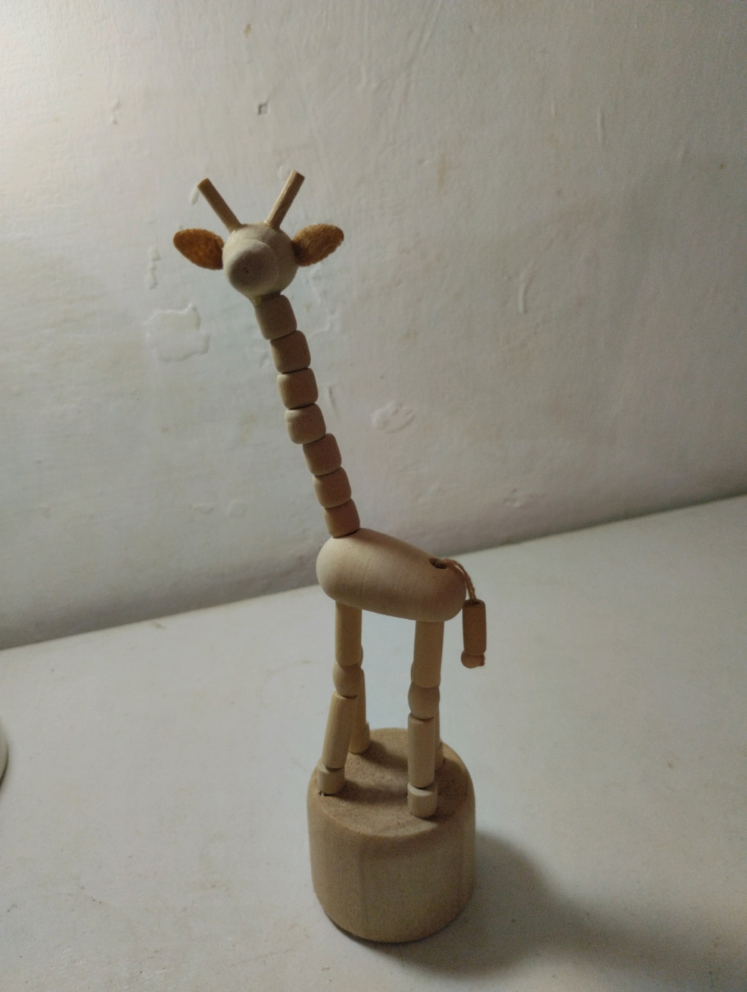 A wooden giraffe figurine stands on a light-colored surface. The figurine has a rounded base, four cylindrical legs, a segmented body and neck, and a head with two small horns and fuzzy ears. A thin string hangs from the back of the giraffe's body. The background is a textured, off-white wall. The scene appears to be indoors, with artificial lighting casting a shadow to the right of the figurine. There is no indication of the location being in Cimenyan, Indonesia, or any specific city.