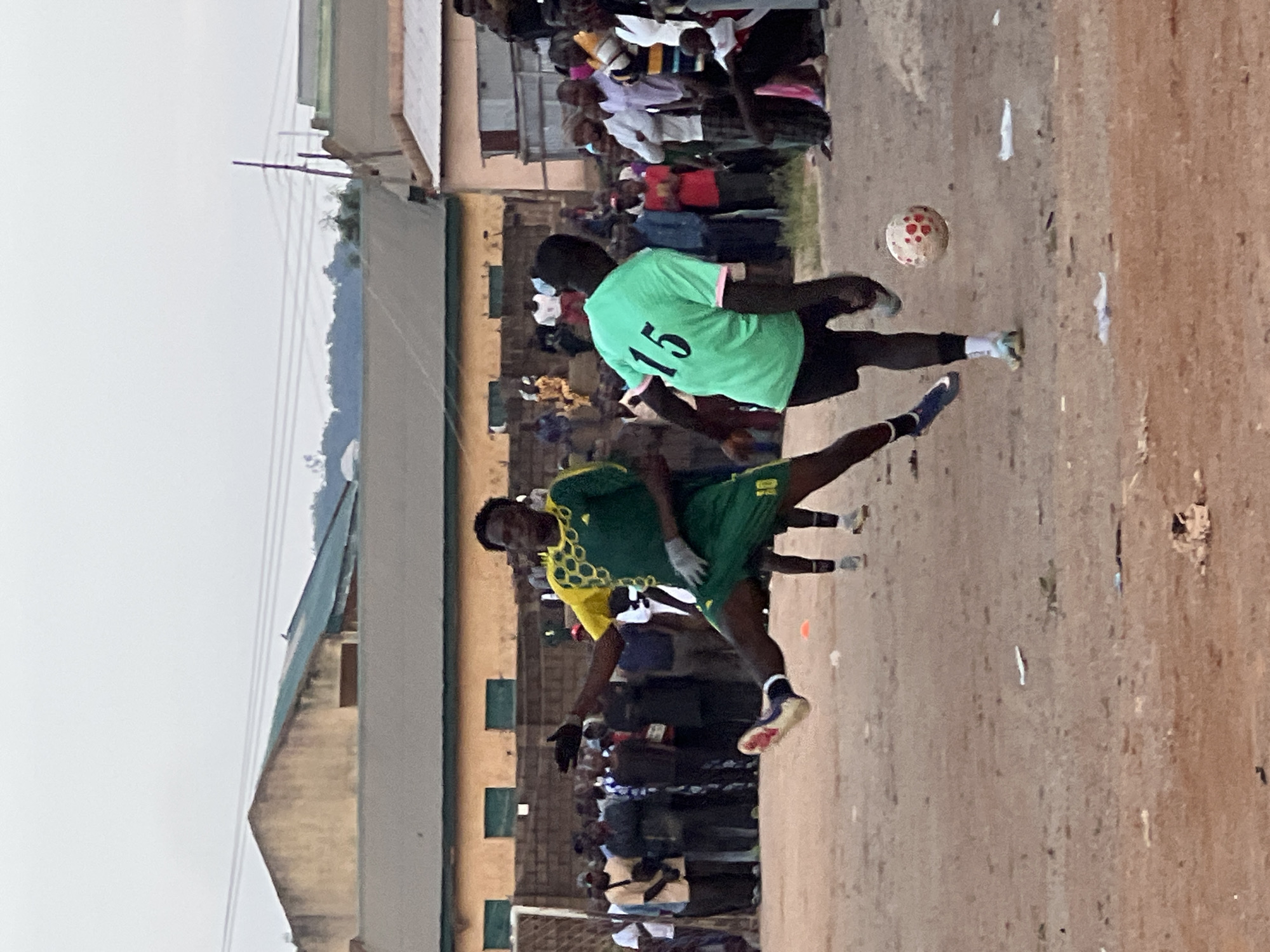 The image depicts a street football match in Jos, Nigeria, with a crowd of spectators gathered in the background. Two young men are actively engaged in the game. One player, wearing a green and yellow jersey with the number 10, is captured in mid-air, kicking a soccer ball with his right leg. He is also wearing gloves. The other player, dressed in a bright green jersey with the number 15 and pink trim, is positioned to receive the ball. The playing surface appears to be dirt or sand, typical of informal street games. The background shows a collection of buildings, including what appears to be a school or community center, and a dense assembly of onlookers observing the match. The sky is overcast, suggesting a late afternoon or early morning setting. The scene conveys a sense of community and lively local sports activity.