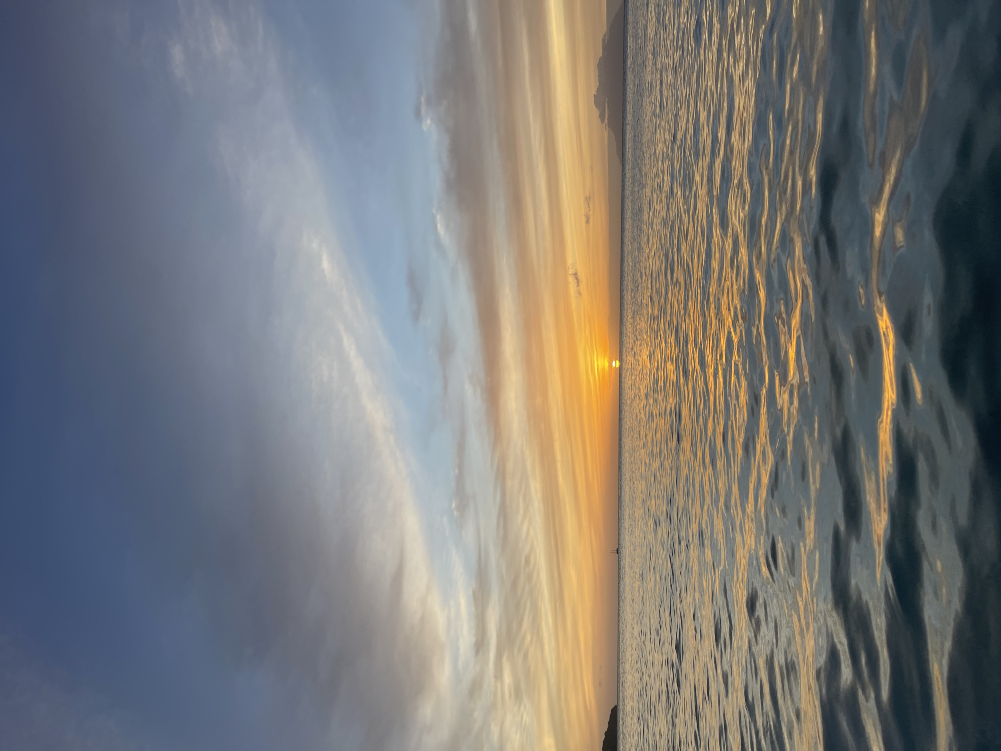 The image depicts a tranquil ocean scene at sunset, with the sun setting over the horizon, casting a warm glow across the sky and water. The sky is a gradient of blue and orange hues, with streaks of clouds illuminated by the setting sun. The ocean is calm, with gentle waves reflecting the colors of the sky. In the distance, a silhouette of a sailboat can be seen on the water, and to the far right, a landmass with a cloudy peak is visible. The overall impression is one of peace and natural beauty. The location is Mare Anglaise, Seychelles.