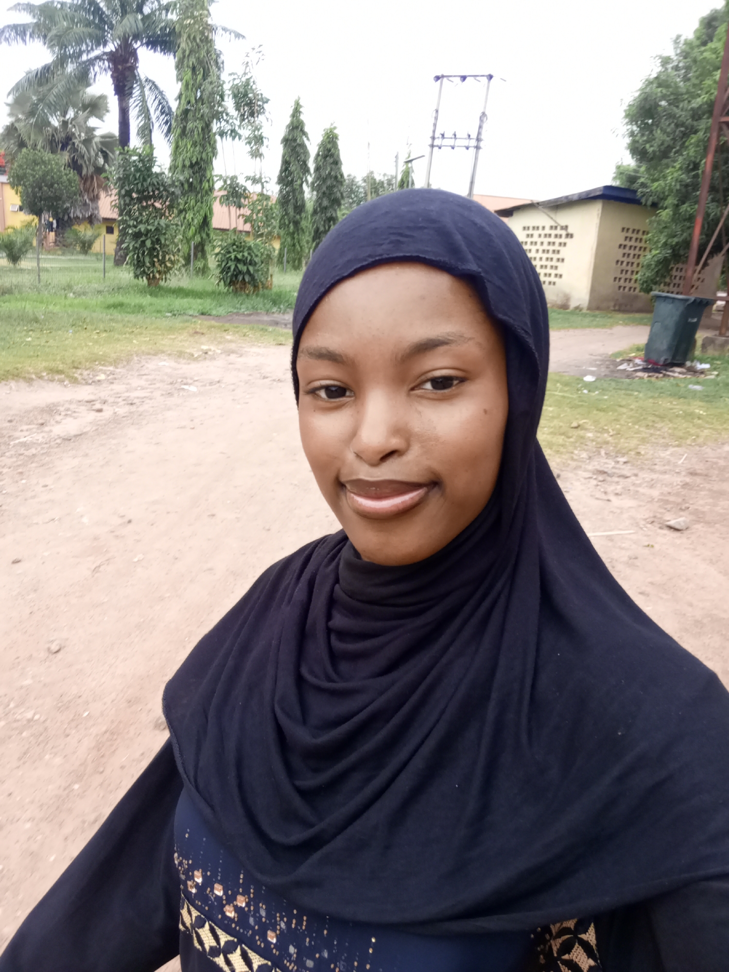 A young woman wearing a black hijab is posing for a selfie in an outdoor setting. The location appears to be in Keffi, Nigeria, judging by the typical building designs and local vegetation visible in the background. She is standing on a dirt or gravel path. Behind her are various trees and buildings, including a light-colored building with a textured exterior wall and a small building with a blue roof. Power lines are also visible. There's also what appears to be a green trash can. The weather is bright but possibly overcast, casting soft lighting on the scene.