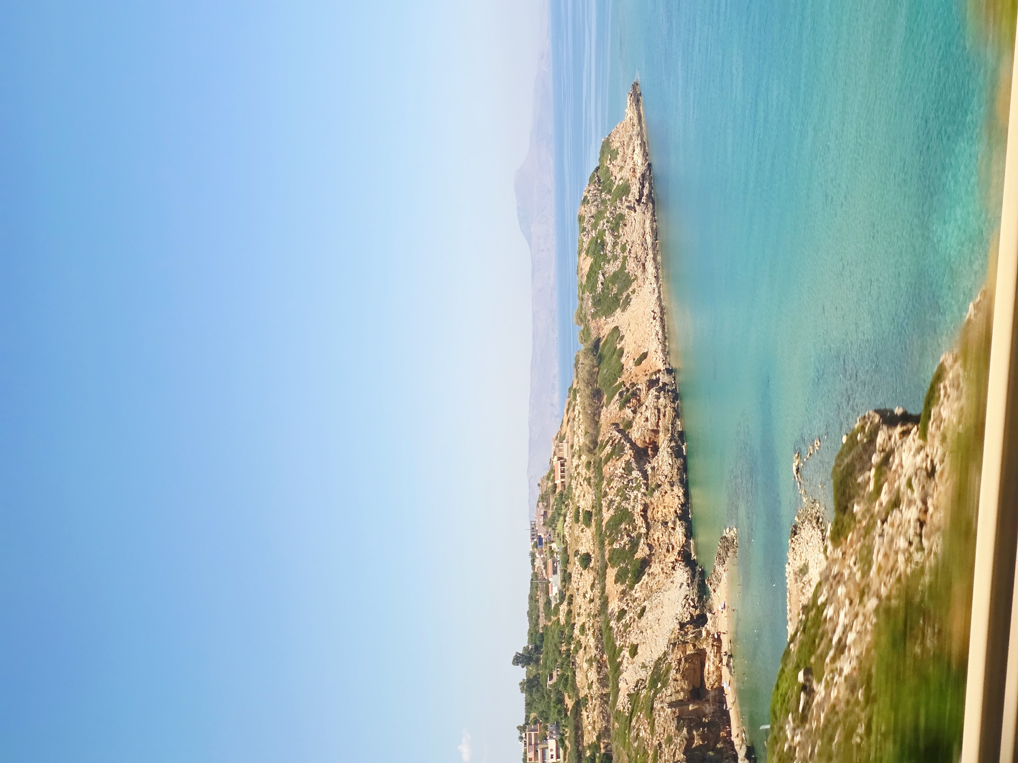 This is a photograph taken from an elevated viewpoint overlooking a coastal scene in Gerani, Greece. The sky is a clear, pale blue, occupying the upper half of the frame. Below the sky, a rugged, rocky coastline extends into the turquoise waters of the sea. The land is covered with dry, sparse vegetation, with scattered trees and bushes. Several buildings, appearing to be houses and villas, are nestled on the hillsides overlooking the coast. The sea is calm and a vibrant shade of turquoise, with the water appearing clear enough to see the seabed in some shallower areas. In the distance, a hazy mountain range is visible. The lower part of the image shows a blurred foreground, suggesting the photo was taken from a moving vehicle, with a glimpse of a sandy or gravelly surface in front of a blurred railing. There are no visible people in the image.