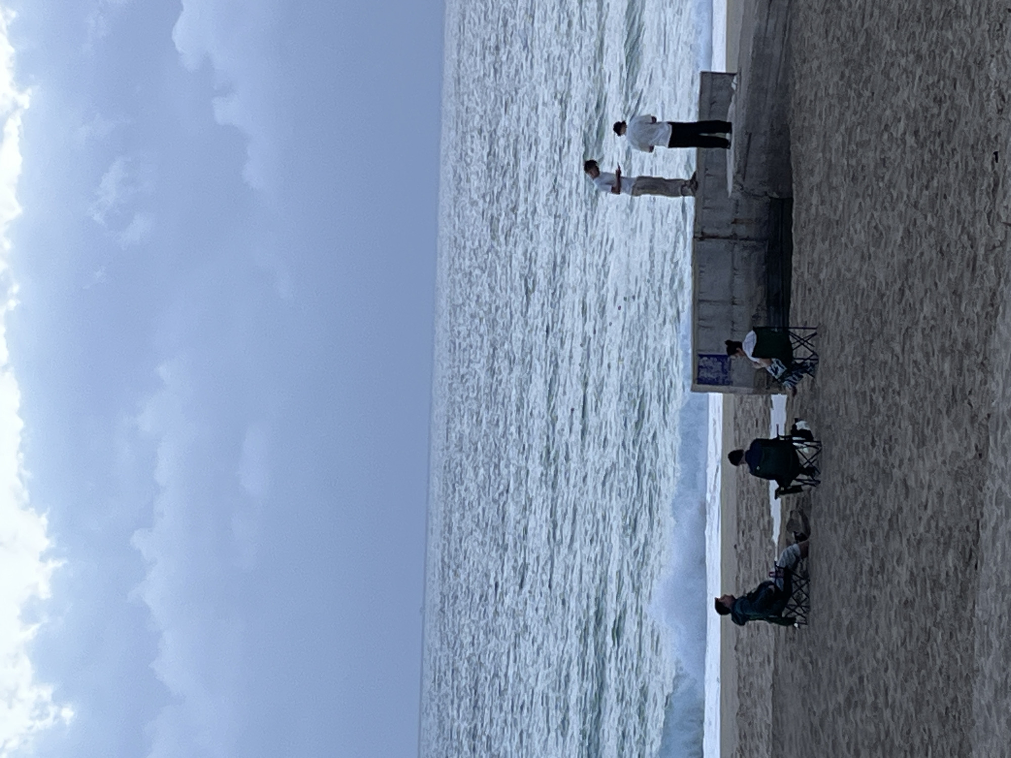 The scene depicts a beachfront in Alanya, Turkey. Two individuals stand on a concrete barrier overlooking the ocean, appearing to observe the waves. Below them, three people are seated in beach chairs on the sand, seemingly relaxing and watching the sea. The sky is overcast with clouds. The environment is a mix of sand, water, concrete, and cloudy sky.