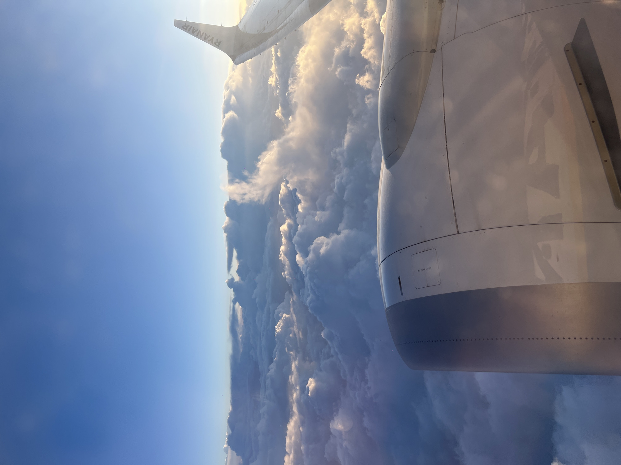 The image shows a view from an airplane window, with a Ryanair winglet visible in the upper left corner and a large jet engine cowling dominating the right side of the frame. Below, a thick layer of clouds stretches across the horizon. The sky above the clouds transitions from blue to a lighter shade, suggesting the time of day might be morning or late afternoon. The scene does not depict any elements that confirm the location in the city of Erkheim, Germany.