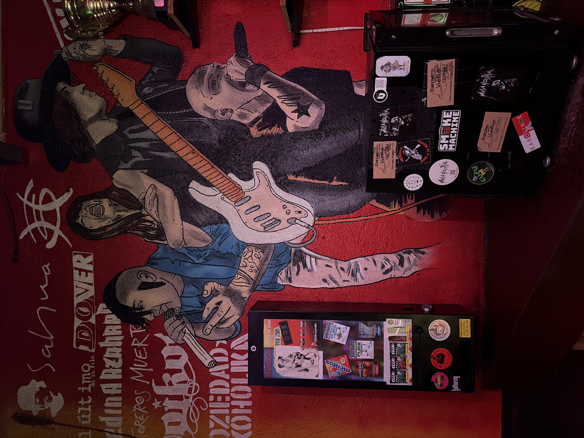 Here is a description of the image:
The photograph shows a section of a wall, likely inside a bar, covered with a mural. The mural features stylized, somewhat graphic drawings of people, possibly musicians or cultural figures, along with names of bands or phrases in what appears to be Spanish. The color scheme is predominantly red with black and white details.
Attached to the wall are several objects: a cigarette/condom vending machine stocked with various products, and above it, a black case or cabinet with numerous stickers affixed to its surface. These stickers display various logos, band names, and other designs. The location is likely Barcelona, Spain, given the presence of the word "SOLIBRA CATALUNYA" on one of the stickers.