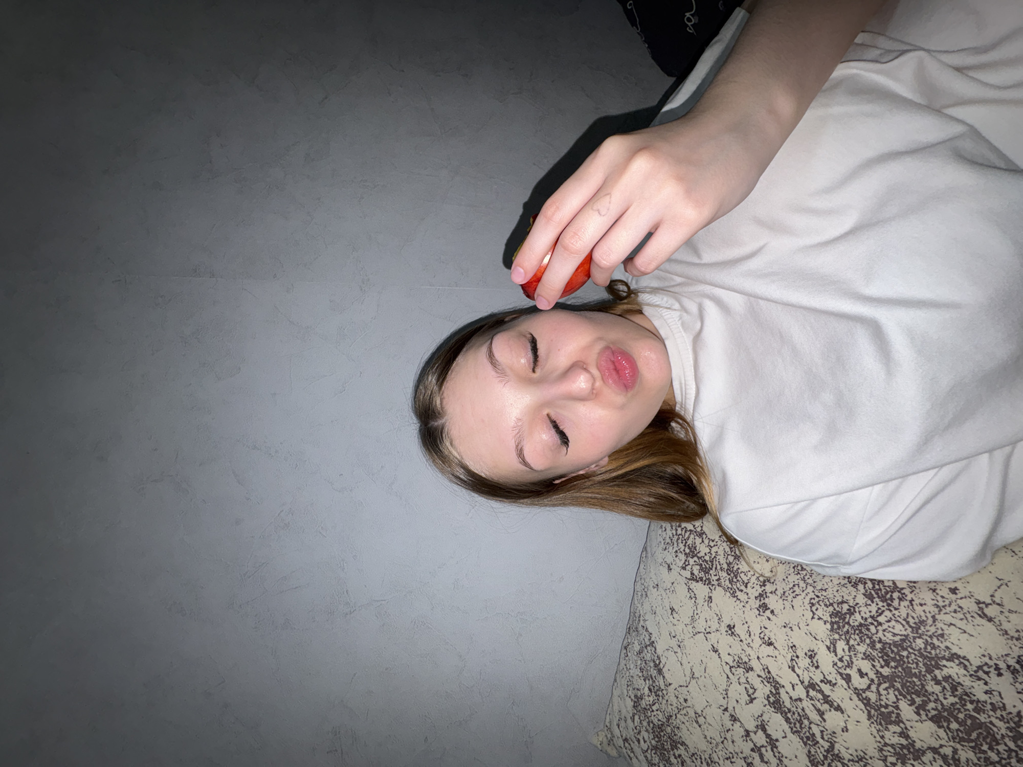 A young woman is lying down, possibly on a bed or cushion, in what appears to be an indoor environment. She is wearing a white shirt and has brown hair. Her eyes are closed, and she is pouting her lips. She is holding a small, red fruit (possibly an apple or tomato) near her forehead. The background consists of a gray wall on the left side and a patterned surface on the bottom right.
There is no information to indicate the location being Chaikovsky, Russia.