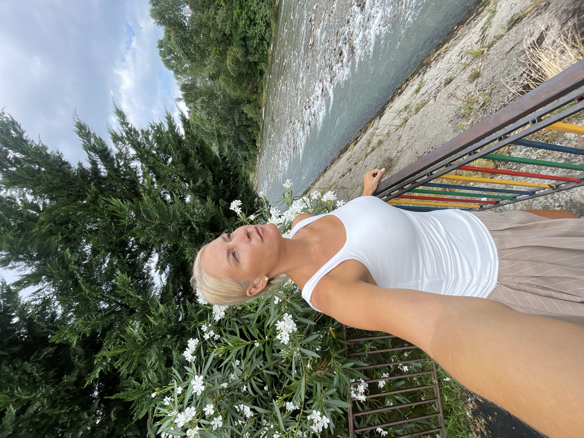 A blonde woman with fair skin is taking a selfie in front of a river in Sochi, Russia. She is wearing a white tank top and a beige pleated skirt. She has her right hand on a railing that is painted with red, yellow, green, and blue bars. Behind her, there is a bush with white flowers, a large green tree, and the river flows through a rocky bed. The sky is overcast.