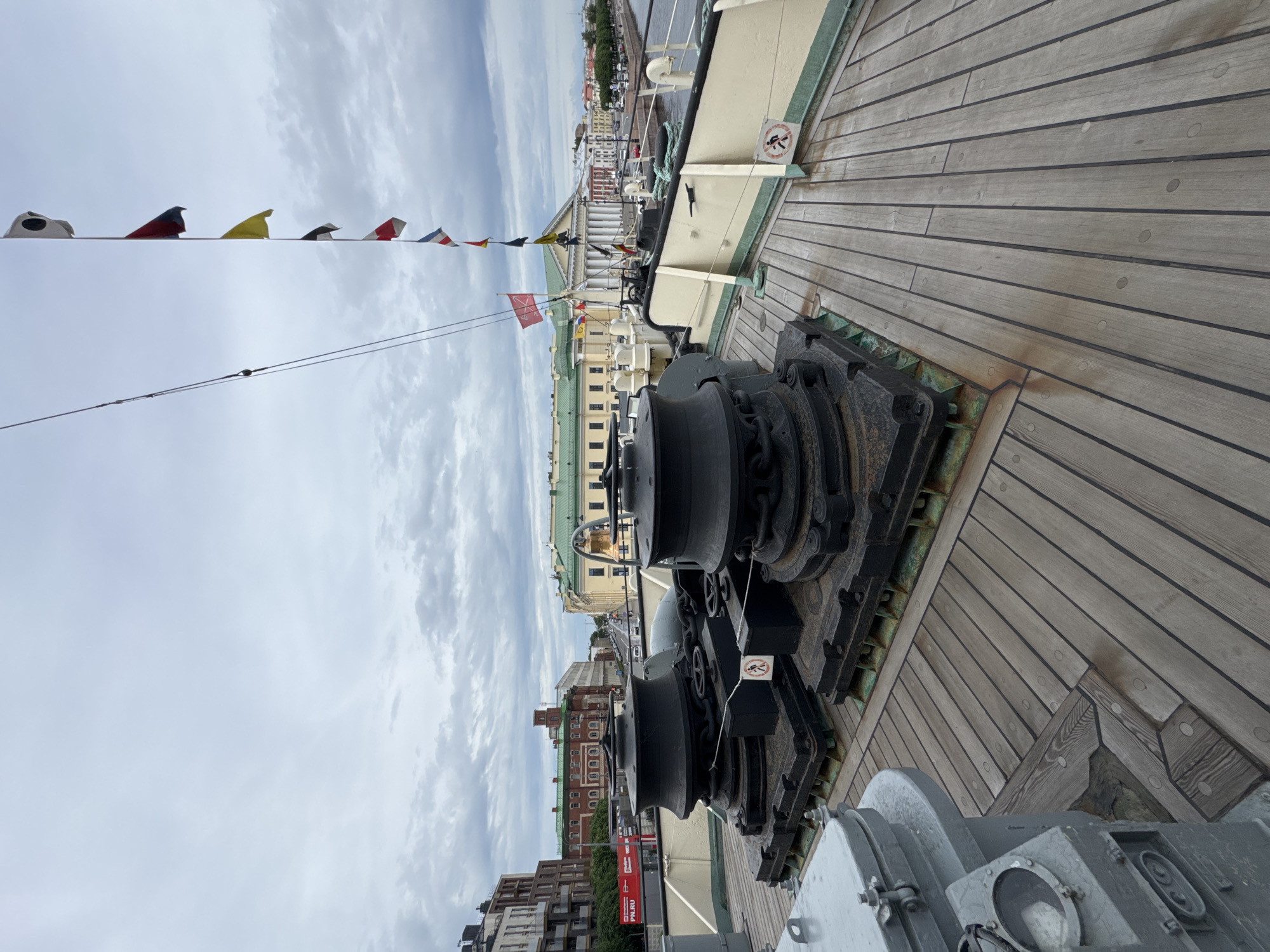 The image shows the deck of a historic ship, possibly a museum ship, docked in Saint Petersburg, Russia. In the foreground are two large, black capstans, which are machinery used for winding rope or cable. The deck itself is made of weathered wooden planks. In the background, a city skyline is visible, with buildings in various architectural styles, including a prominent building with a classical facade and a large green dome. A body of water, likely a canal or river, separates the ship from the city. Numerous flags, including nautical signal flags and what appears to be a red ensign, are strung along a mast. The sky is overcast, suggesting a cloudy day. The overall scene depicts a maritime heritage site within an urban environment.