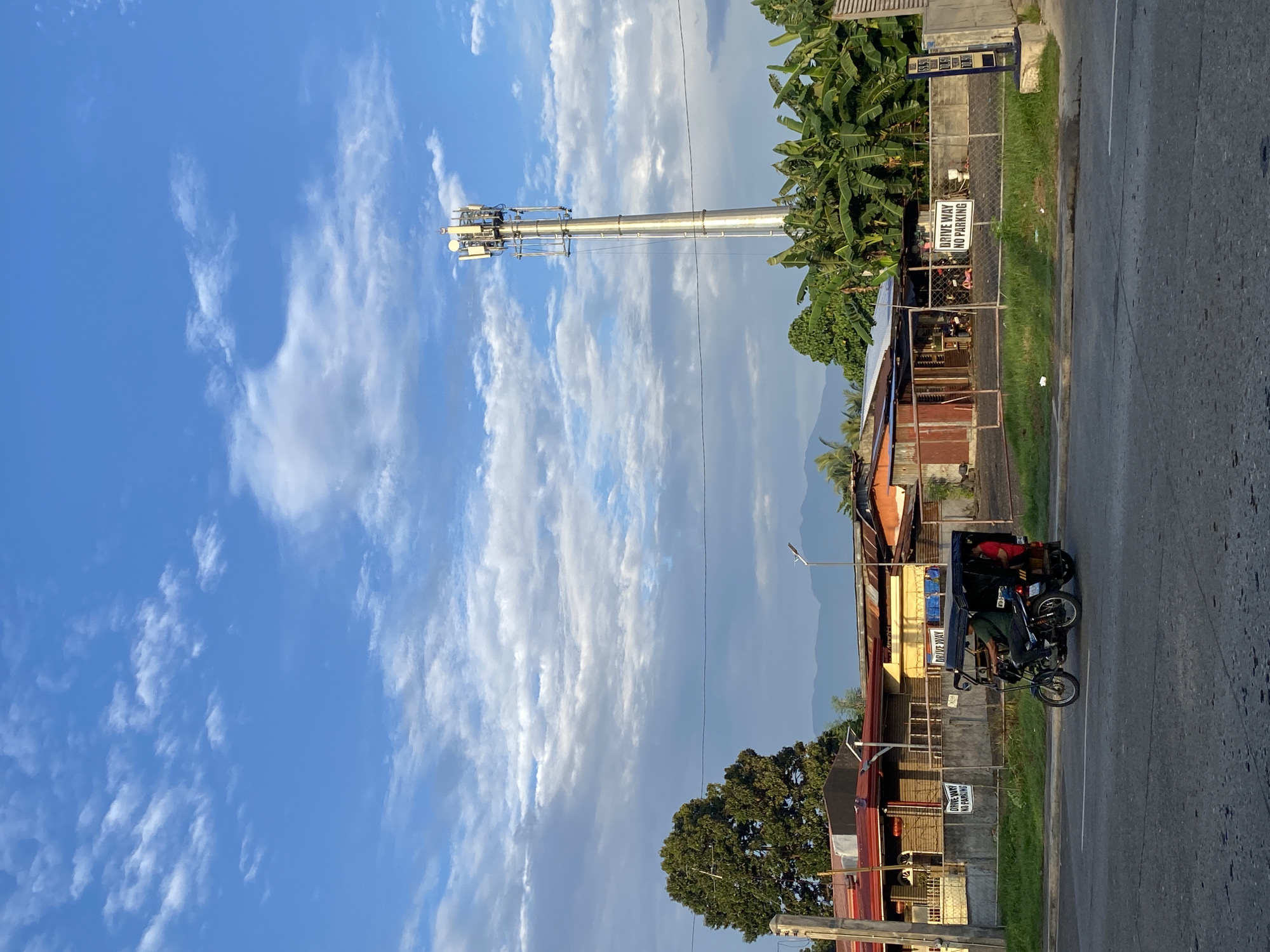 This is a medium-shot, vertical photograph of a street scene in Davao City, Philippines. The sky is a vibrant blue, with scattered white clouds. A tall cell tower dominates the right side of the frame, topped with antennas and equipment. In the middle ground, a row of single-story buildings with various roofing materials and structures, including what appears to be a sari-sari store, is visible. A chain-link fence separates these buildings from the road. Two "DRIVE WAY NO PARKING" signs are posted on the fence. A motorela, a three-wheeled motorized vehicle commonly used as public transport, is moving along the paved road in the foreground. It carries two passengers, a driver and a person in the back. Banana trees and other foliage are abundant in the background, and a mountain range can be seen in the distance. The overall lighting suggests it is either early morning or late afternoon.