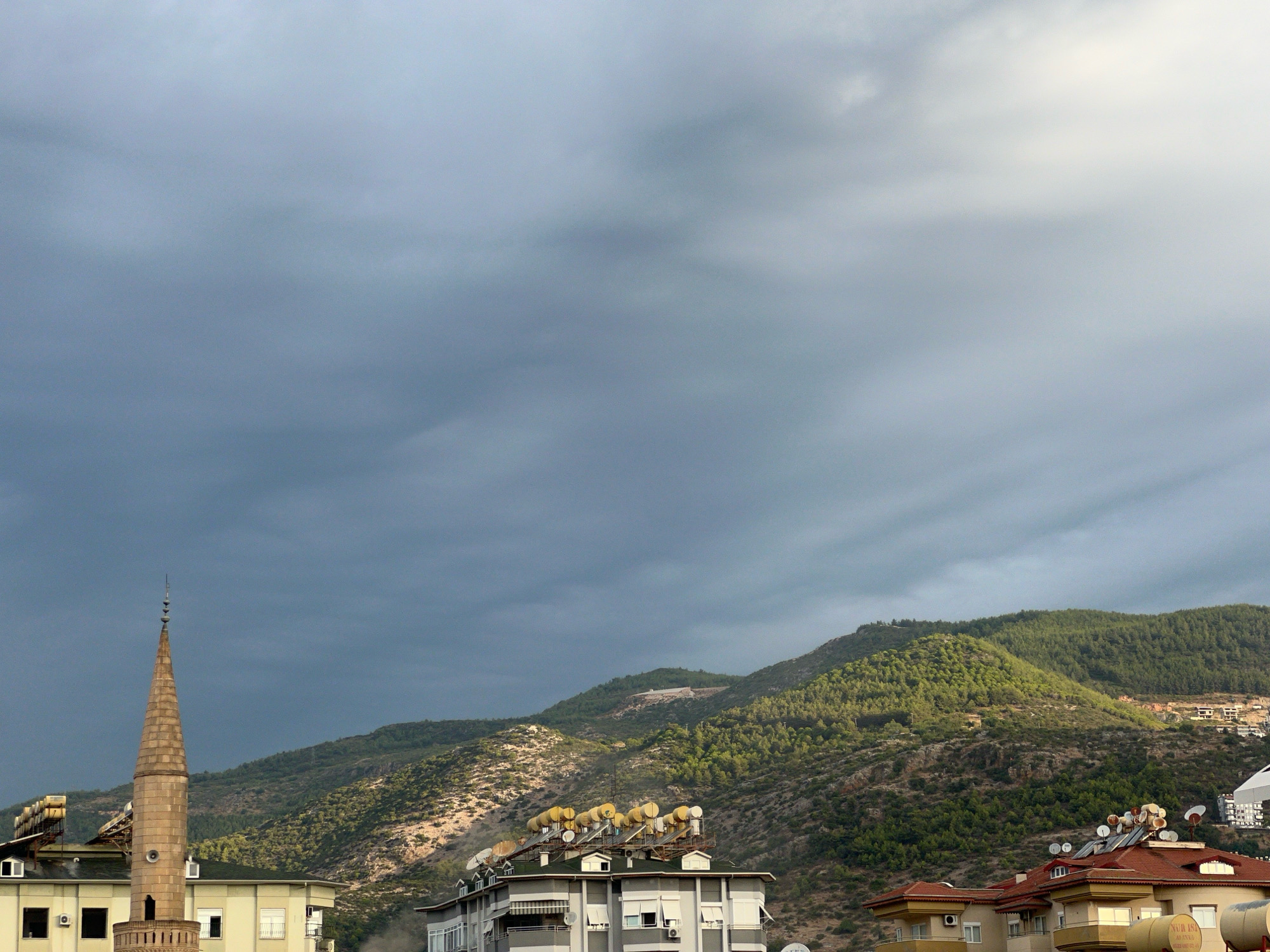 The image depicts a panoramic view of Alanya, Turkey, with a dramatic sky filled with heavy, grey clouds. In the foreground, a tall, slender minaret with a light brown, stone construction stands prominently on the left. It is topped with multiple small decorative elements. Adjacent to the minaret are several buildings, one with a cream-colored facade and visible air conditioning units and windows. Another building to its right features a grey facade with a green roof, adorned with numerous solar water heaters and satellite dishes on its rooftop. Further to the right, a building with a red-tiled roof is partially visible, also equipped with satellite dishes. In the background, lush green, tree-covered hills rise, with some rocky, barren slopes interspersed. The hills are dotted with distant houses and buildings, suggesting the urban sprawl extending into the natural landscape. The overall atmosphere is subdued, likely due to the overcast weather. No people are visible in the scene.