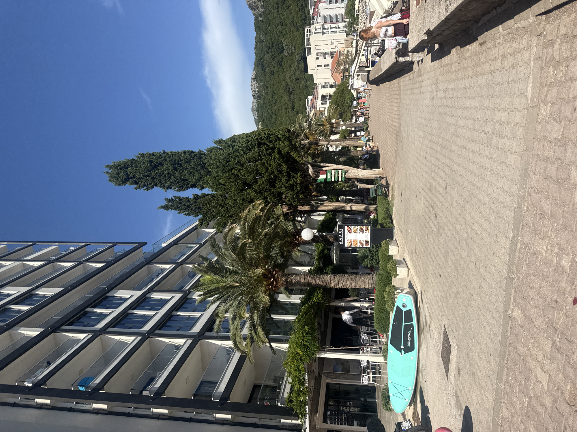 The image depicts a sunny day in a coastal town, likely Rafailovići, Montenegro. The scene features a modern hotel building with numerous balconies, palm trees, and other lush greenery. In the foreground, a turquoise stand-up paddleboard rests on a paved walkway, suggesting recreational activities are popular in the area.
Beyond the hotel, a tree-lined promenade is visible, with several people strolling or sitting at outdoor cafe tables. Further in the background, white buildings climb a verdant, mountainous hillside, characteristic of a Mediterranean landscape. The sky is a clear, bright blue with a few wispy white clouds. The overall atmosphere is relaxed and leisurely, indicative of a vacation destination. The time of day appears to be late morning or early afternoon due to the strong sunlight and shadows.
Notable details include a sign with a menu outside one of the establishments, which appears to say "CAPRI". The stand-up paddleboard has the brand name "THUNDER" visible on it. The overall impression is one of a vibrant and attractive tourist location.