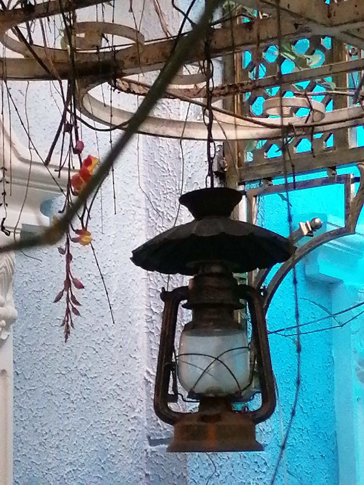 The media file depicts an old-fashioned kerosene lantern hanging from a metal structure, possibly part of an outdoor patio or gazebo. The lantern, made of dark metal with a glass enclosure containing what appears to be a white liquid or substance, hangs by a chain. It is intricately designed, with a decorative top and a sturdy handle.
In the background, a textured, light-colored wall suggests an outdoor setting. Behind the wall, a vibrant blue, possibly stained glass or plastic, forms a backdrop, adding a splash of color to the scene. This blue element is framed by a decorative metal lattice structure, hinting at an architectural feature.
Hanging to the left of the lantern are delicate vines with small, reddish-orange flowers and buds. These organic elements provide a contrast to the man-made objects and add a touch of natural beauty. The overall lighting suggests it might be daytime, though the light quality is somewhat diffused.
The scene evokes a sense of nostalgia and rustic charm, possibly set in a garden or a quaint establishment. The combination of the antique lantern, the delicate flowers, and the ornate metalwork creates a visually interesting composition. The location context of Da Lat, Vietnam, a city known for its French colonial architecture and cool climate, further supports the imagery of a potentially charming and slightly melancholic atmosphere. No visible text is present in the image.