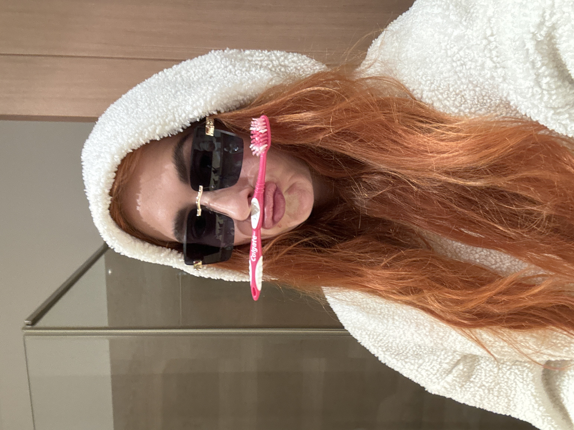 A woman with red hair is lying down, wearing a white hooded robe and black sunglasses. She is holding a pink Colgate toothbrush in front of her face. The background appears to be a bathroom with a cabinet. The location in Budva, Montenegro cannot be determined from the image.