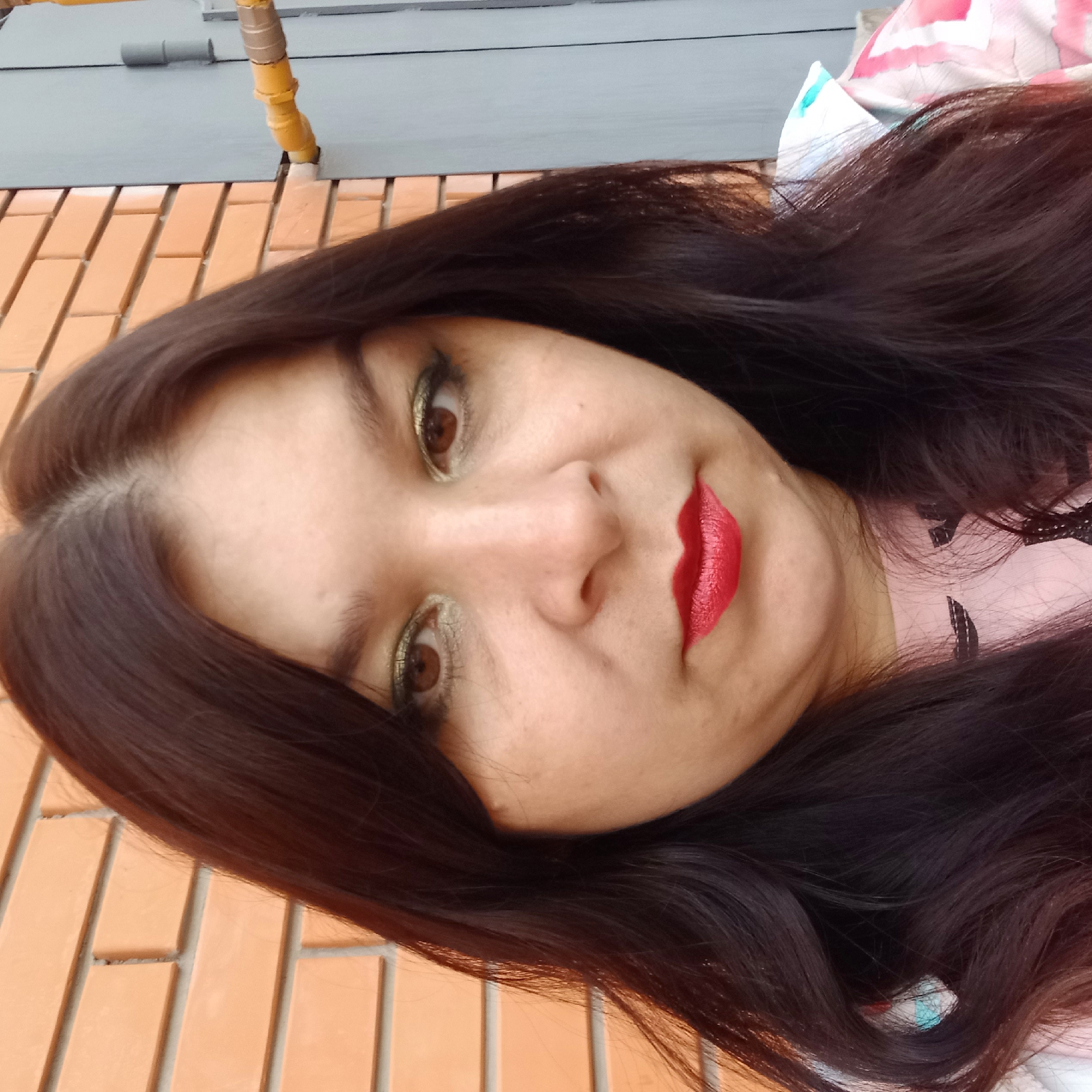 In Kropyvnytskyi, Ukraine, a medium-shot, eye-level selfie features a woman with dark brown hair, styled with a center part and loose waves. She is wearing bold makeup, including shimmery green eyeshadow and bright red lipstick. The woman is positioned in front of an orange brick wall on the left and a gray metal door with yellow pipes on the right. Her face is the central focus of the image, with her expression neutral. She is wearing a pink shirt with a graphic print. The lighting appears to be natural daylight.