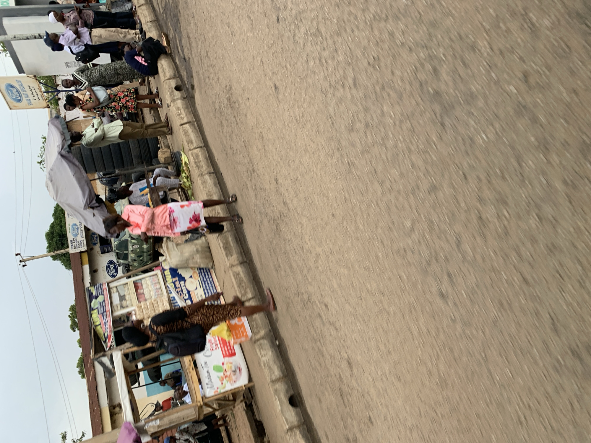 The image depicts a street scene in Rafin Par, Nigeria. A group of people stand near a sidewalk adjacent to a road. Several businesses are visible, including a Ford spare parts shop and a food stall selling fruit salad and corn. People are engaged in conversation and standing on the sidewalk next to the road. The road is paved.