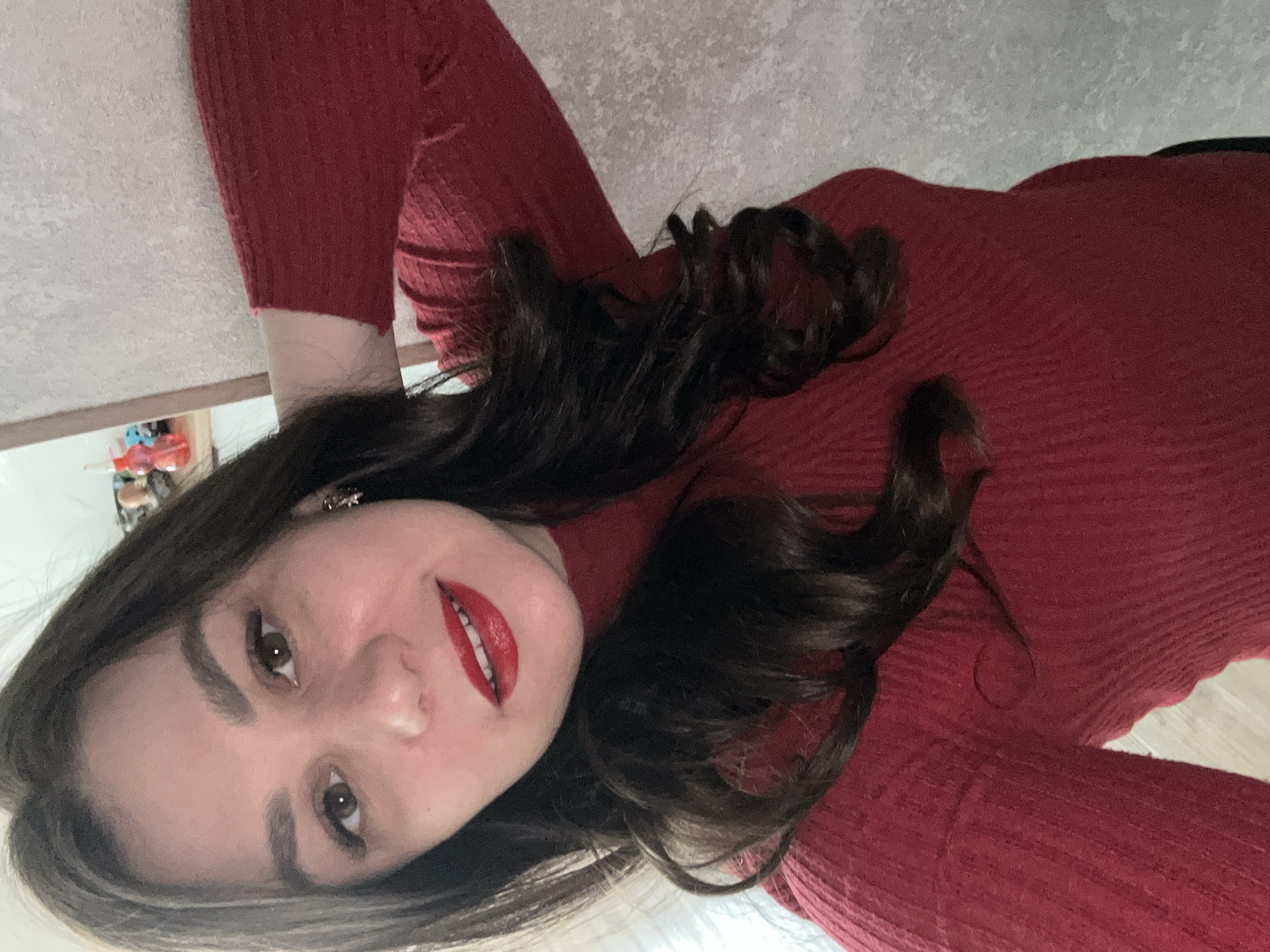 A person is wearing a red, ribbed long-sleeved garment. The person has dark brown hair that is curled and styled. The person has fair skin, brown eyes, and is wearing red lipstick. A blurred background appears to show a wall, and a shelf containing small objects is visible in the top left of the frame. The location is unknown and cannot be determined based on the image. The city of Chaikovsky, Russia, cannot be confirmed.