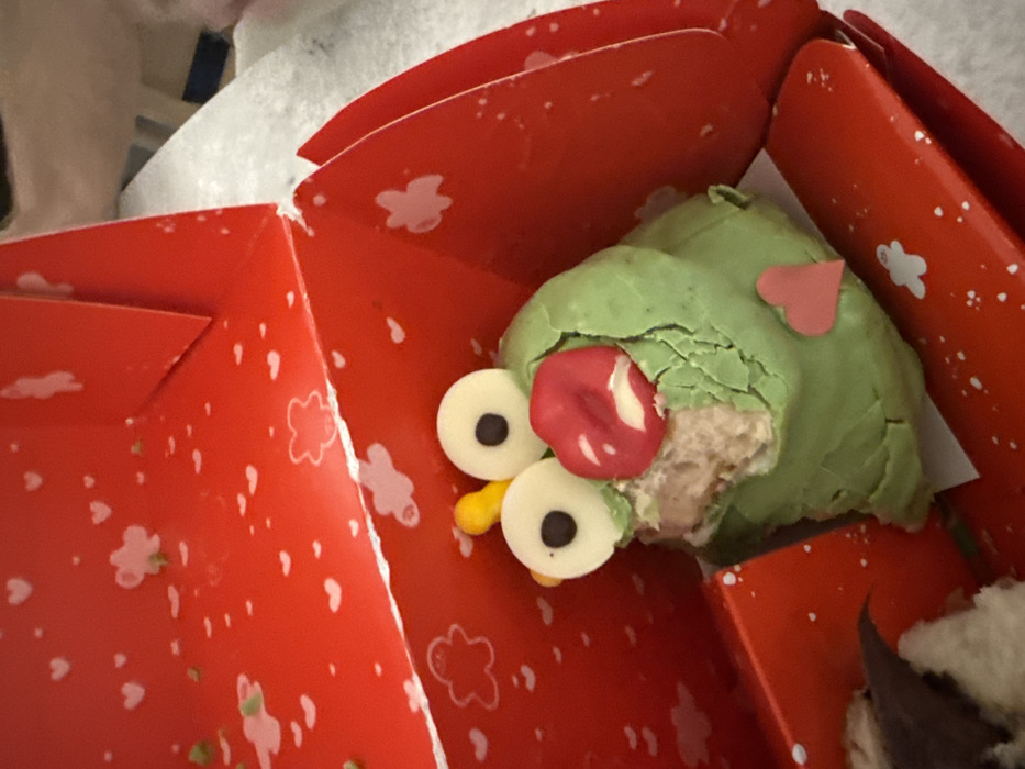 A close-up, high-angle shot depicts a partially consumed confectionery item contained within an open red cardboard box. The confectionery is approximately ovoid, primarily green, and decorated to resemble a whimsical character. It features two prominent white circular eyes with central black pupils, a set of red, pouted lips serving as a mouth, and a small yellow, conical protrusion above its right eye. A small pink heart-shaped decoration is affixed to the lower front-right side of the green exterior. A significant portion of the confectionery has been removed from the front-center, exposing a light brown, crumbly interior, consistent with cake or a similar baked good. The green exterior appears to be a thin coating that has cracked where bitten. The red cardboard box is patterned with numerous small white hearts and larger, abstract white cloud or flower shapes. A corner of the box is visible, showing its folded construction. In the lower-left corner of the box, a blurred, brown and white object, possibly another confectionery item, is partially visible. The immediate background is a uniformly soft, out-of-focus white surface, likely a table or countertop. The scene is situated in Barcelona, Spain.