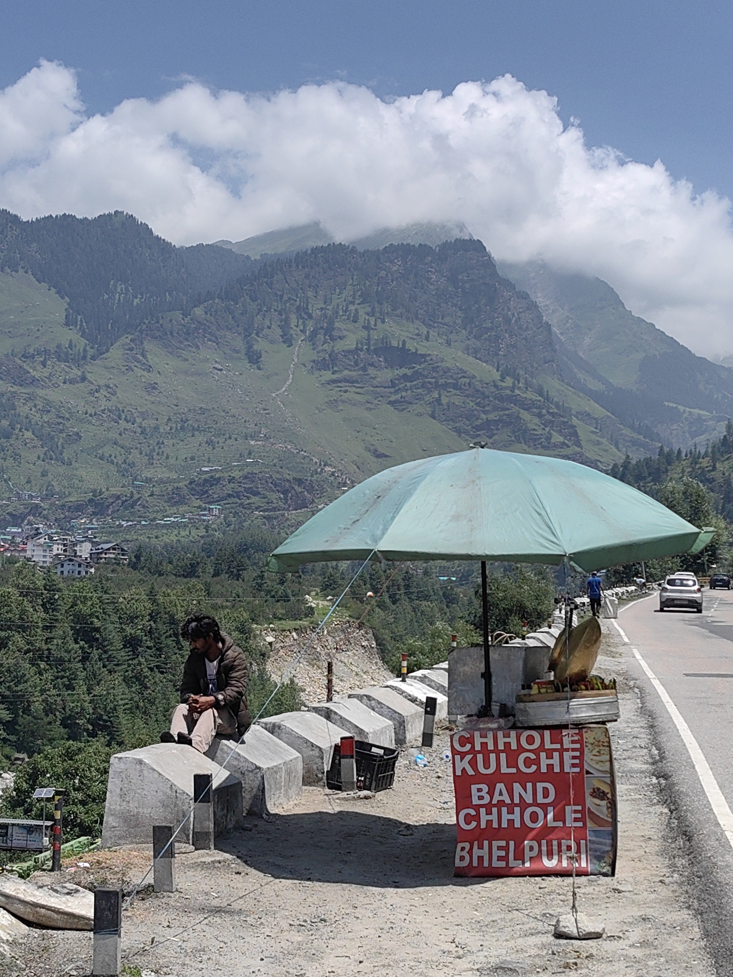 The image shows a roadside scene with a food stall in front of a mountain range. A man is sitting on a concrete barrier facing away from the camera. He is wearing a brown jacket and tan pants. A large, light-colored umbrella is providing shade for the food stall, which has a red sign that reads "CHHOLE KULCHE BAND CHHOLE BHELPURI." There are various food items displayed on the stall. A road runs along the right side of the image, with a white car and a black car driving on it. The background features lush green mountains with some buildings visible in the distance. The sky is blue with scattered white clouds. The scene appears to be located in Kulang, India.
