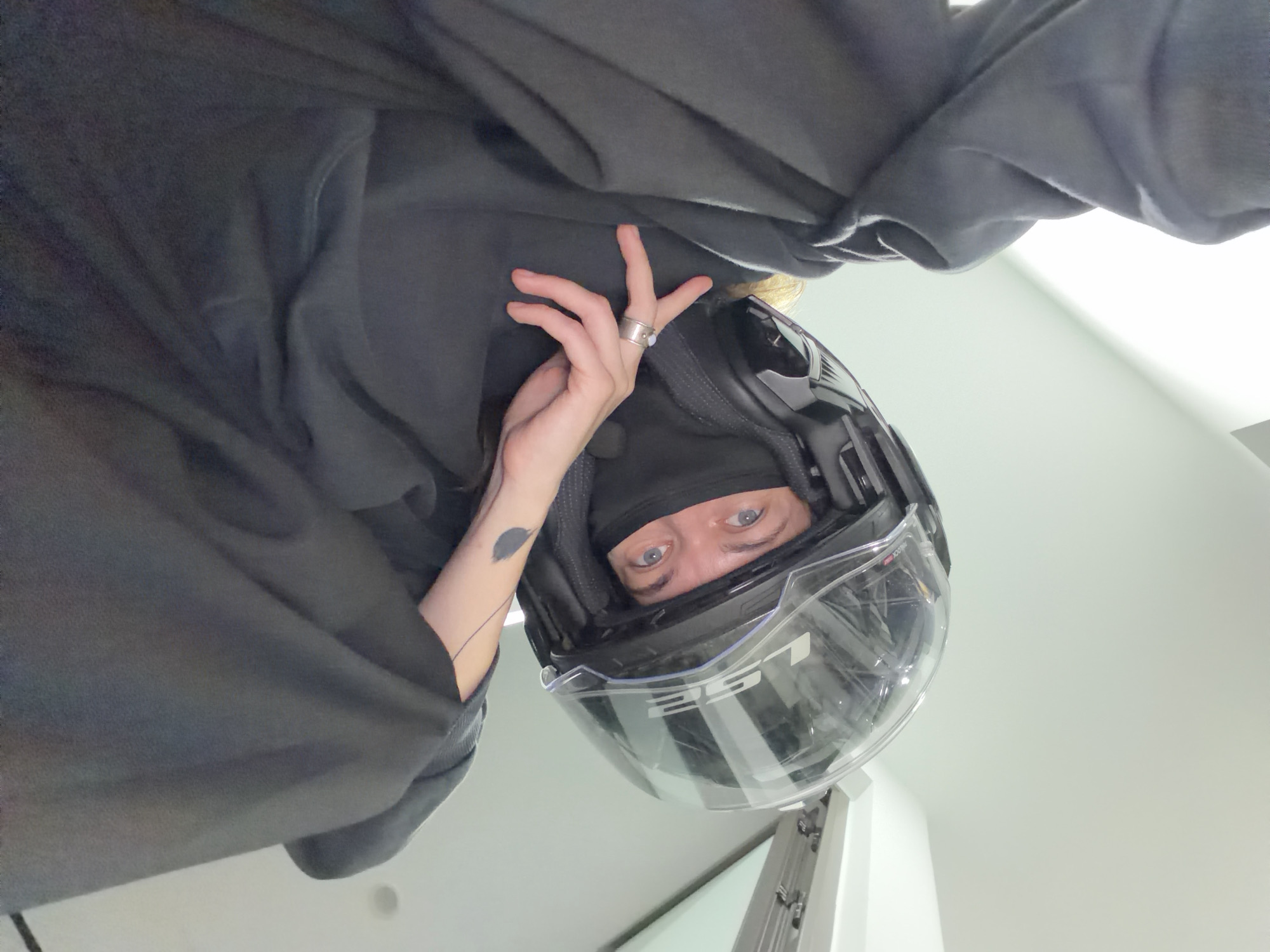 A person is depicted wearing a full-face motorcycle helmet and a black balaclava, with only their eyes visible. They are also wearing a black hooded garment. A ring is visible on their hand, and a tattoo is visible on their forearm. The photograph appears to have been taken from a low angle, looking up at the subject. The background is a plain, light-colored wall. There is no indication of the location being Đà Nẵng, Vietnam, or any other city.