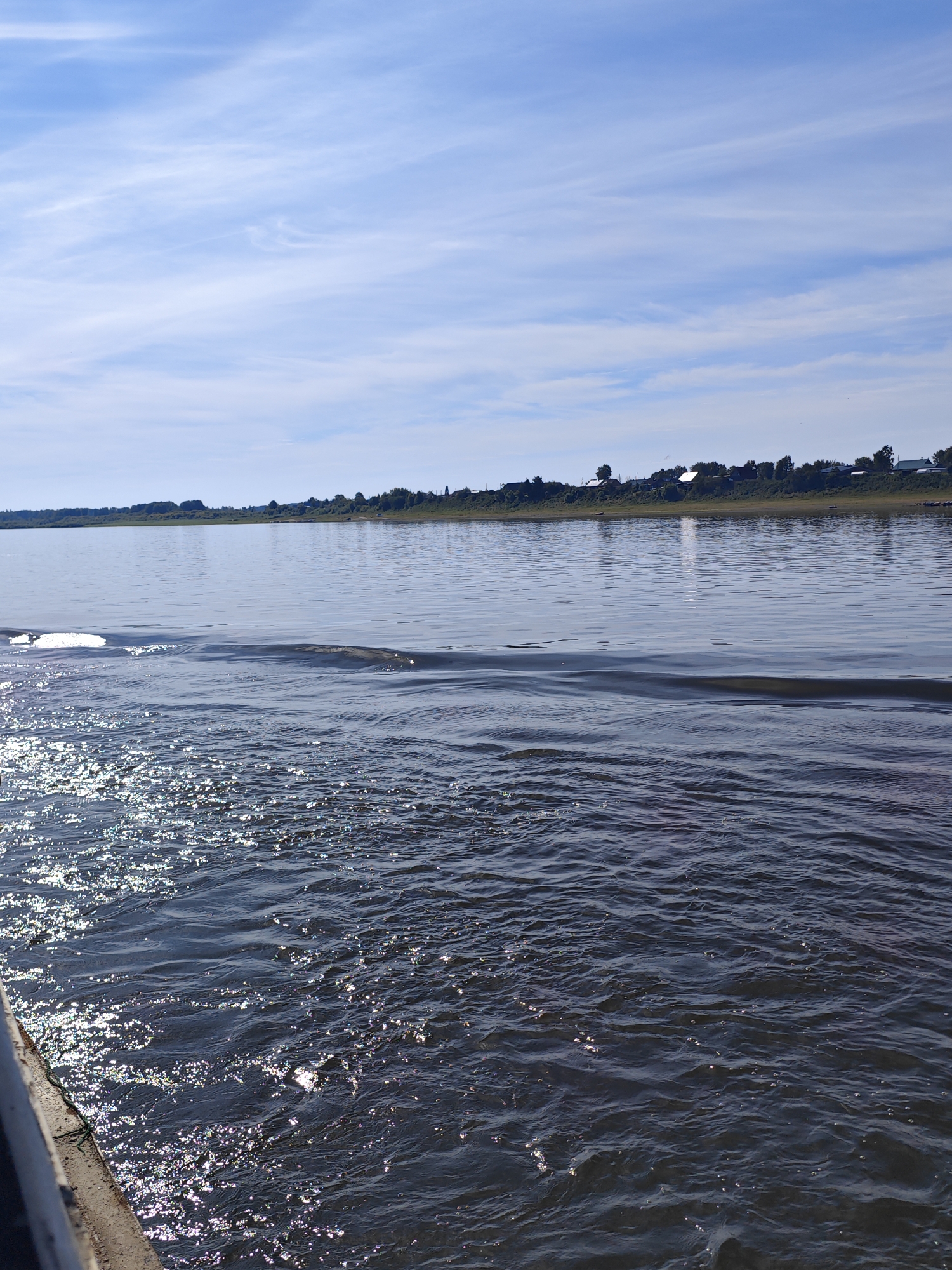 The image shows a wide, calm river on a sunny day. The water is dark and reflects the blue sky with thin white clouds. On the left side of the image, the water is rippled and shows a bright reflection of the sun. A concrete embankment is visible in the bottom left corner. In the distance, across the river, is a shoreline with a line of trees and houses. The sky is a clear blue with wispy white clouds. The scene appears to be a tranquil natural landscape, likely near a settlement given the houses visible. This scene is consistent with the appearance of the Dnieper River as it flows through or near the city of Yartsevo, Russia, a city located on the Dnieper.