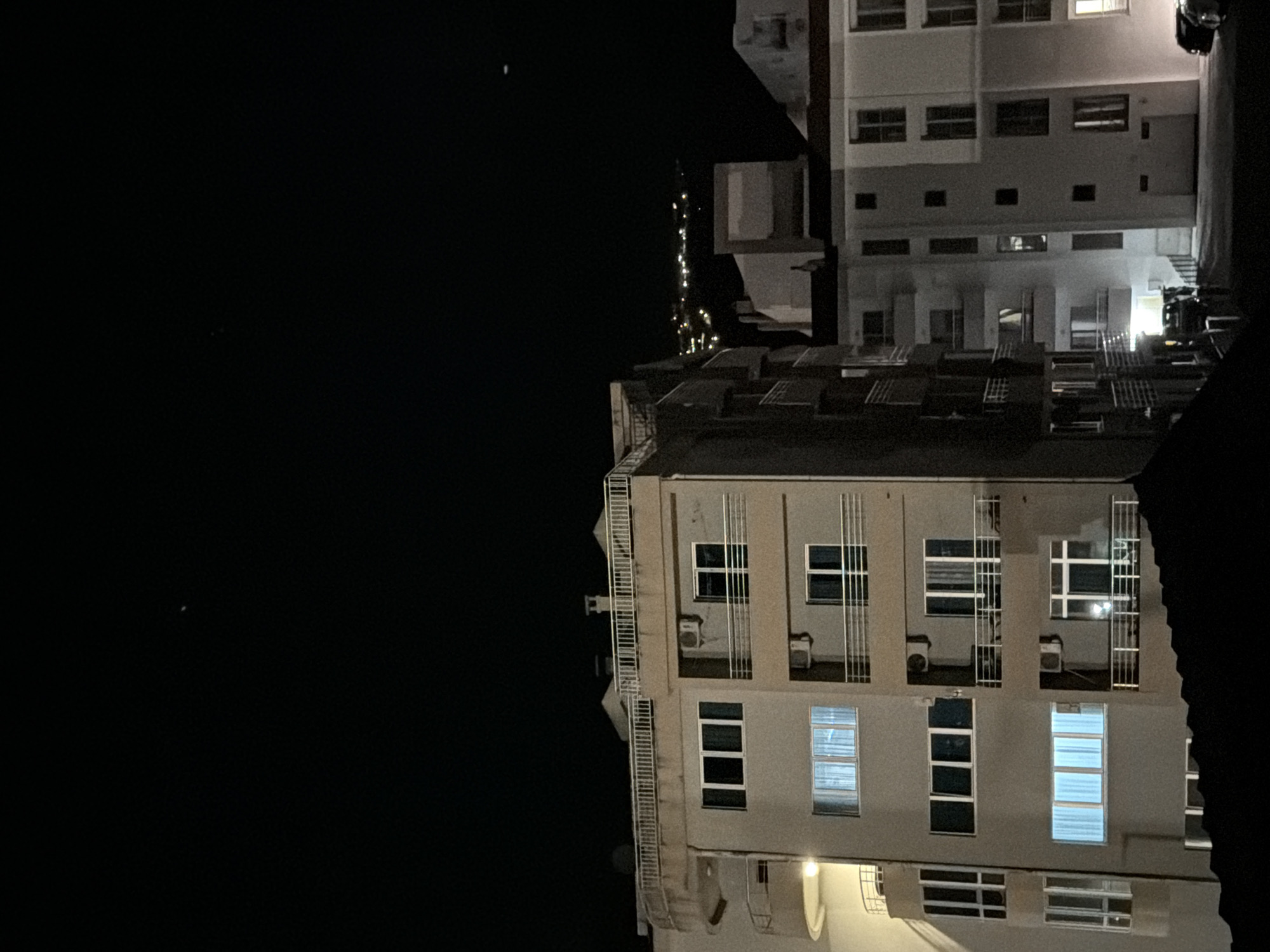 A nighttime image shows a building with multiple stories in Budva, Montenegro. The building is illuminated by the light from its windows. A fire escape is on the side of the building. The sky is dark. There is another building in the background.