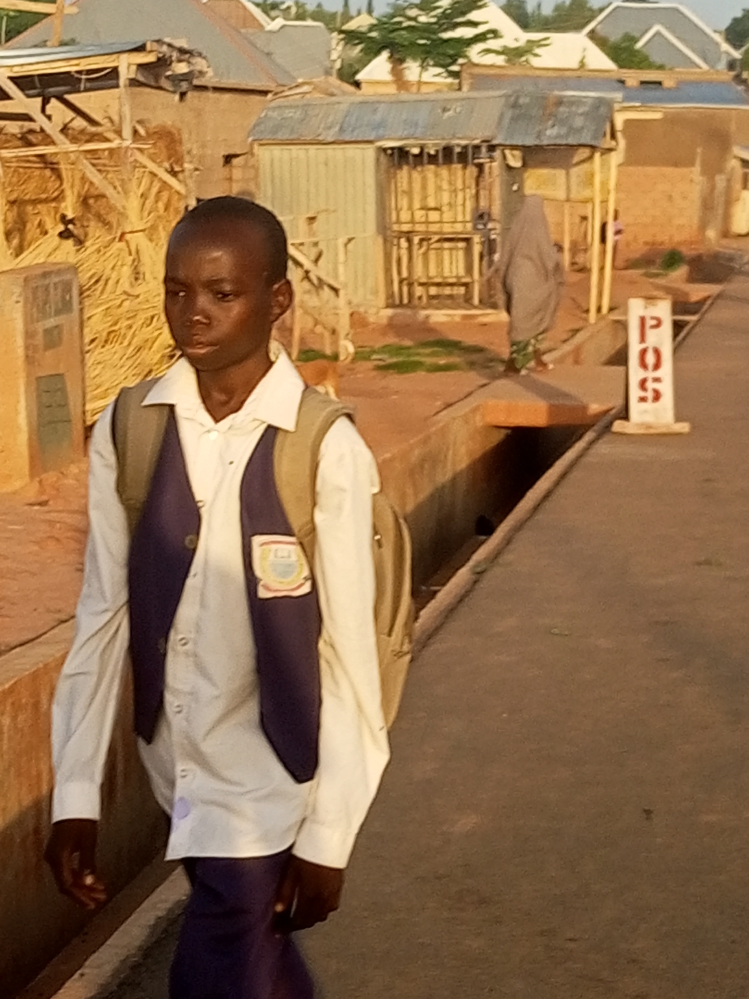A young boy walks on a dirt road in a dusty, urban environment, likely in Tempure, Nigeria. The boy, wearing a white collared shirt, a dark purple vest, and dark pants, carries a light brown backpack. He has dark skin and short, dark hair. In the background, a woman covered in a grey hijab walks away from the camera. Various structures are visible, including buildings with corrugated metal roofs, some appearing to be makeshift stalls constructed with wood and straw. A white sign with red lettering that reads "POS" stands to the right of the frame, indicating a point-of-sale terminal. The ground is unpaved, and a drainage ditch runs alongside the road. The lighting suggests it is late afternoon or early evening.