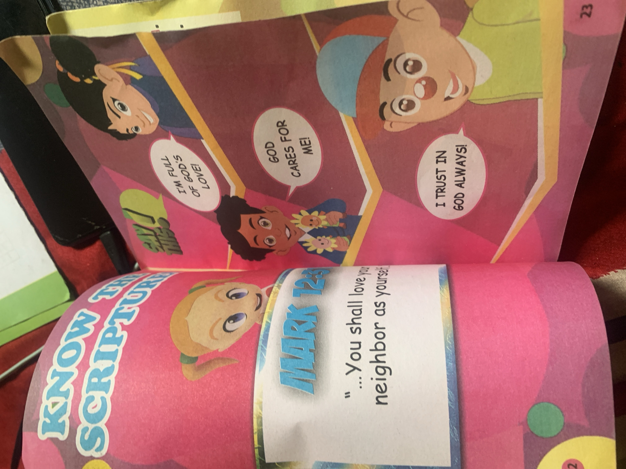The image displays an open children's book with colorful cartoon illustrations and text. On the left page, large blue text reads "KNOW THE SCRIPTURE" in a curved font, followed by the text "MARK 12:31" in blue block letters. Below this, in black text, is a quote: "...You shall love your neighbor as yourself." A cartoon girl with blonde pigtails is visible in the upper left.
The right page features three cartoon children in speech bubbles. The top right child, with dark hair in buns and a purple shirt, has a speech bubble saying "I'M FULL OF GOD'S LOVE!". The middle child, with dark curly hair and a blue shirt, holds a flower and has a speech bubble that says "GOD CARES FOR ME!". The bottom right child, wearing a green shirt and a blue and orange hat, has a speech bubble that reads "I TRUST IN GOD ALWAYS!". The page is bordered with a yellow and orange stripe. The number "23" is present in the bottom right corner. The background appears to be a dark red, textured material, likely the binding or a table. The image does not contain any elements that would allow for the identification of a specific location within Jauro Yinu, Nigeria.