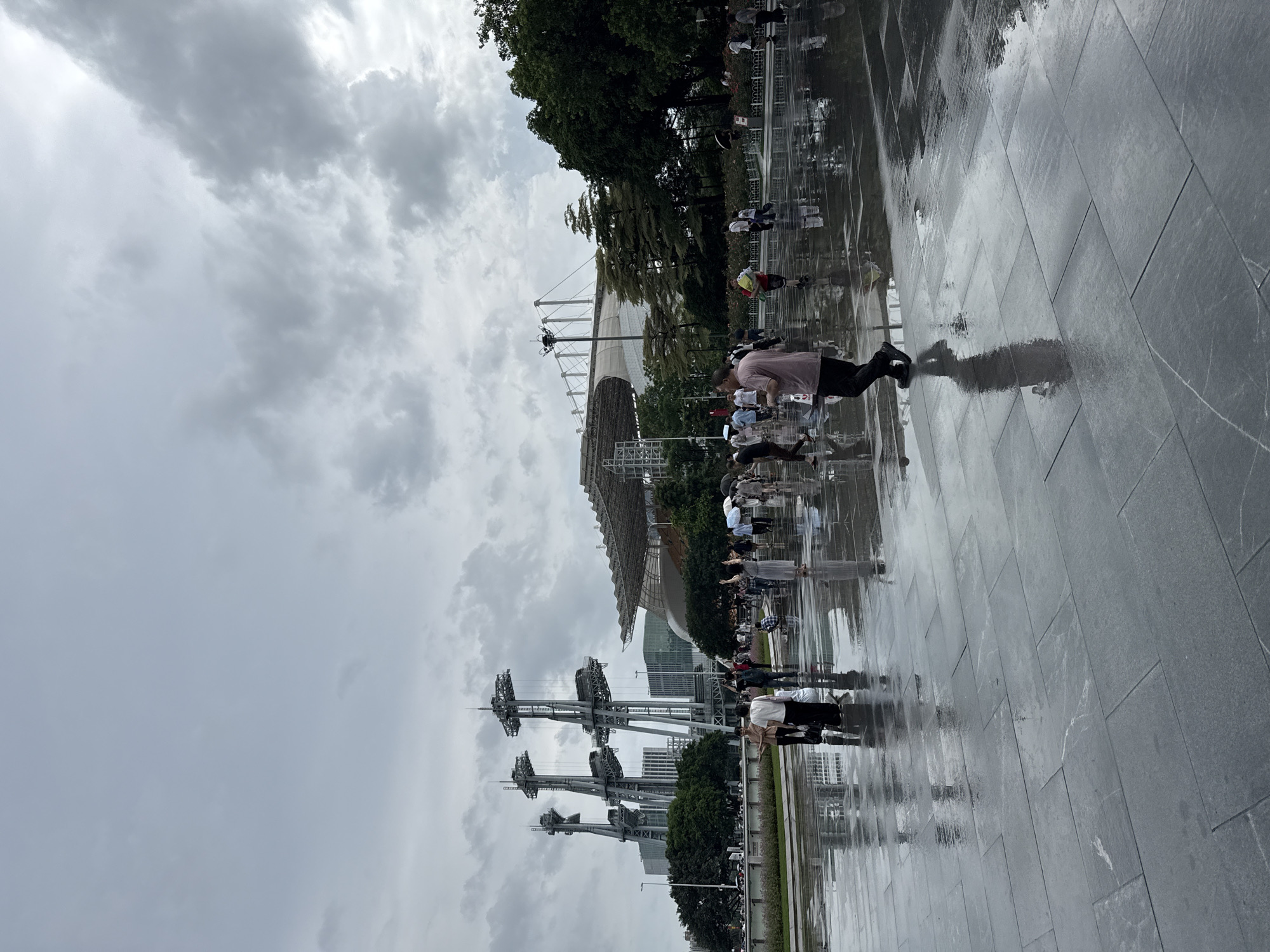 The image captures an outdoor scene under a cloudy sky, possibly in the Tianhe District of China, with a large modern stadium visible in the background. The ground is paved with large, dark stone tiles that are wet and reflective, indicating recent rain or damp conditions. Several people are scattered across the paved area, some walking, others standing and observing. A man in a pinkish shirt and dark pants is walking towards the right side of the frame, his reflection clearly visible on the wet ground. Other individuals are seen in smaller groups or individually, some holding umbrellas, suggesting it might be drizzling or has recently rained. In the mid-ground, there are trees and some modern architectural structures, including tall, metallic towers with platforms. The overall atmosphere appears subdued due to the overcast weather.