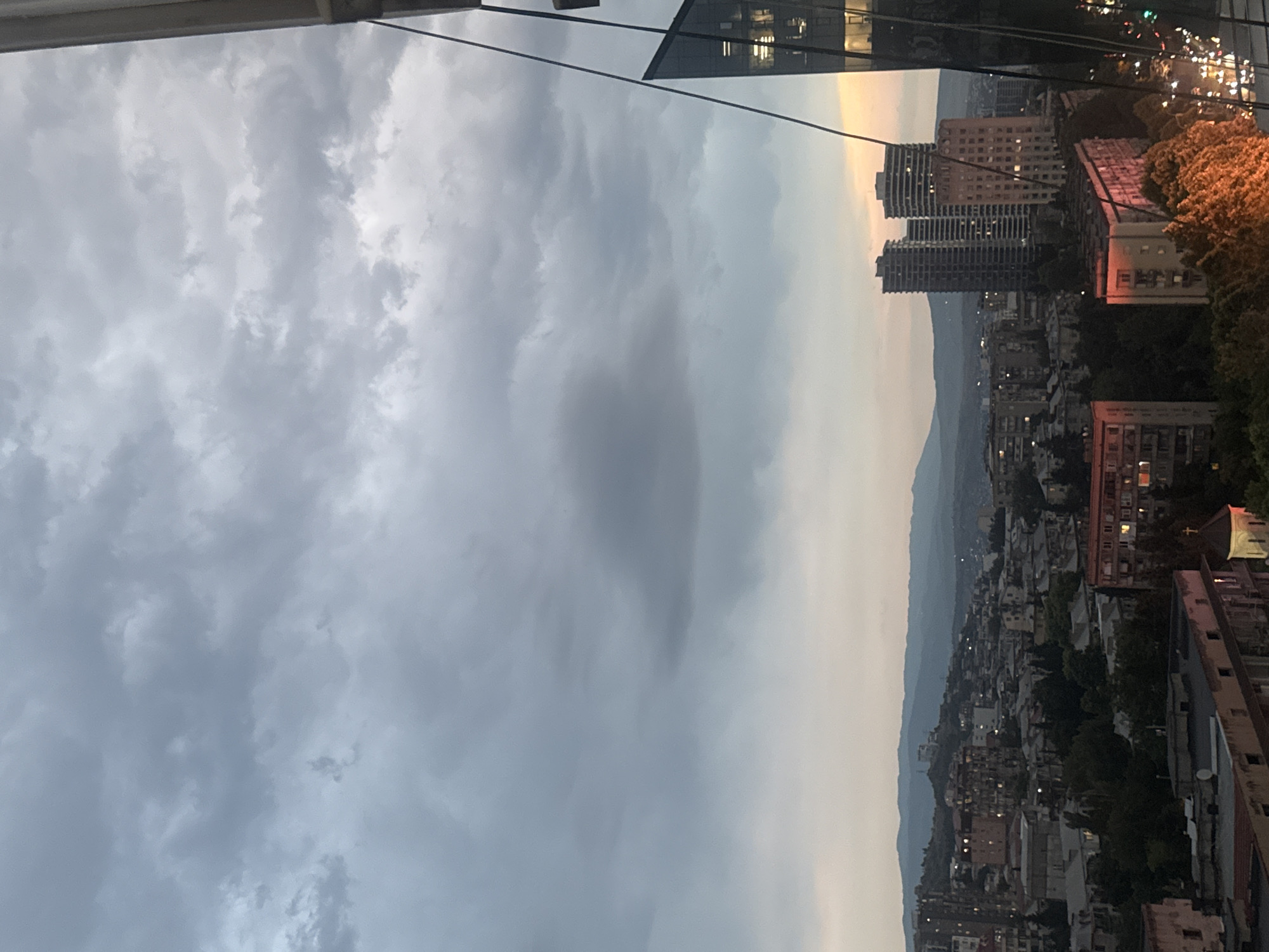 The image captures a panoramic view of Tbilisi, Georgia, at dusk. The sky is filled with dramatic, heavy clouds, casting a moody atmosphere over the cityscape. In the foreground, a modern glass building with illuminated windows is partially visible on the right, its angled facade reflecting the dimming light. Beyond this, the urban landscape unfolds with a dense collection of buildings, ranging from older, mid-rise apartment blocks to more contemporary high-rises. Lights are beginning to twinkle in many of the buildings, indicating the transition from day to night.
The city stretches out towards a distant range of hills and mountains, silhouetted against the twilight sky. Greenery, in the form of trees, is interspersed throughout the urban fabric, particularly noticeable in the lower part of the image where some trees are brightly illuminated by warm light, possibly from streetlights or nearby buildings. A small church with a dome and a cross is visible amongst the buildings in the middle ground, adding a touch of architectural history to the scene. The overall impression is one of an active city settling into the evening under a brooding sky. There are no people or animals visible in the frame.