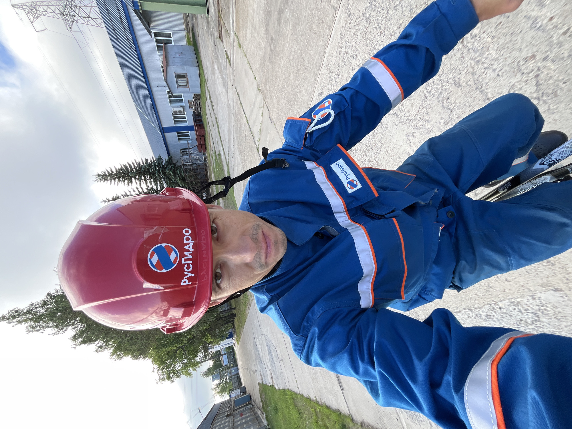 A man wearing a red hard hat and a blue uniform with reflective stripes stands outdoors. The hard hat has a logo that reads "РусГидро" (RusHydro). The uniform also features the RusHydro logo on the sleeve. He appears to be in an industrial or utility area, with buildings and power lines visible in the background. The location is identified as the city of Chaikovsky, Russia.