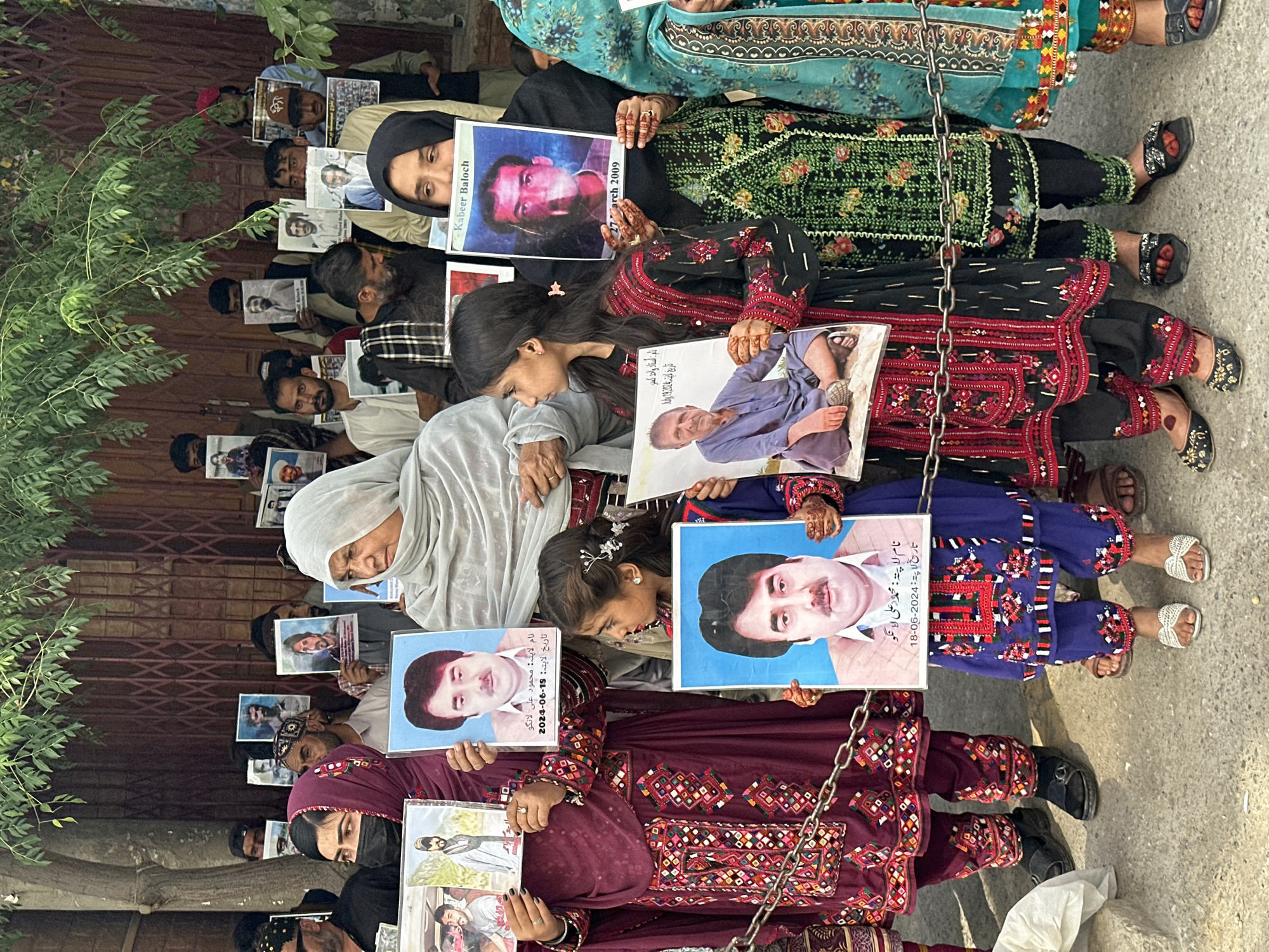 A gathering of people in Quetta City Tehsil, Pakistan, are holding photographs of individuals, possibly in a protest or memorial. Some people sit in the front, dressed in traditional clothing, while people wearing headscarves are seated behind them. Each person holds a photograph of someone. Captions under the photographs state name and date of disappearance.