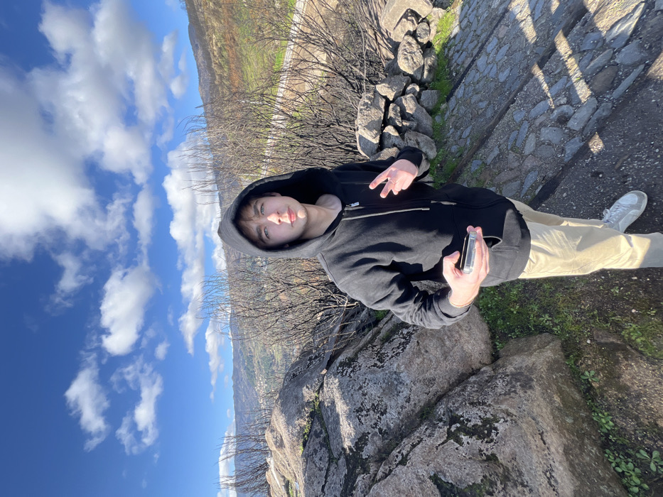 An outdoor, wide-angle photograph taken in a mountainous region identified as Vila Real, Portugal, depicts a single male subject. The male, appearing young adult, is centrally positioned, wearing a black hooded zippered sweatshirt with the hood up, light-colored khaki trousers, and white athletic shoes. He holds a smartphone in a clear case in his left hand at waist level. His right hand is raised, displaying a two-finger "peace sign." His gaze is directed towards the camera with a neutral expression.
The foreground consists of a cobbled path underfoot, bordered by large, moss-covered boulders and sparse green vegetation to the left, and a low stone wall constructed from stacked rocks to the right. The mid-ground features numerous leafless, dark, slender shrubs and branches, consistent with dormant or fire-affected vegetation. The background reveals a valley containing a small town and undulating hills with varied green and darker tones, under a clear blue sky with scattered white clouds. A winding road is visible on the distant hillside. The scene is illuminated by direct sunlight.