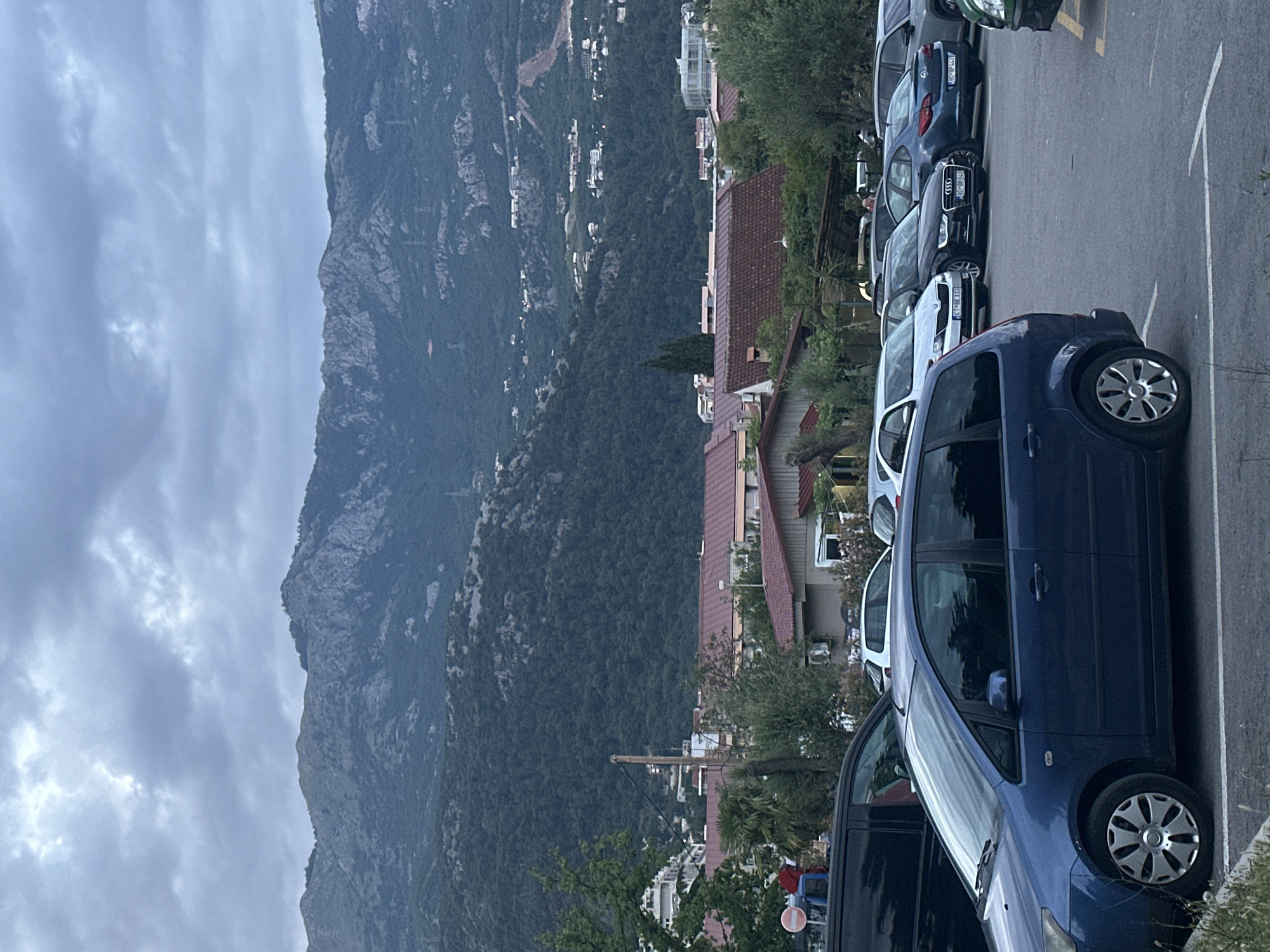 The image depicts a mountainous landscape and residential area in Budva, Montenegro, under an overcast sky. In the immediate foreground, a paved parking lot is visible, with a dark blue Ford C-Max minivan parked prominently on the left. Behind it, a black van is partially visible. Further into the parking lot, other parked vehicles include a white Skoda Rapid (license plate CT BE 333), a black Audi (license plate RO BB 848), a dark blue Opel Astra (license plate RO BP 443), and several other cars of varying colors.
The midground features numerous residential buildings with distinct red-tiled roofs, nestled among abundant green trees and shrubs, including a palm tree. A wooden utility pole stands centrally on the left side of the midground, with overhead lines extending from it. A circular red and white sign is partially visible on the far left.
The background is dominated by extensive, dark green forested mountains with rugged, rocky peaks, stretching across the horizon. Small, scattered white structures are discernible on the lower slopes of these mountains, indicating distant human settlements. The sky is entirely covered by dense, grey clouds, suggesting an overcast day or impending weather change. No specific individuals or discernible actions are present within the frame.