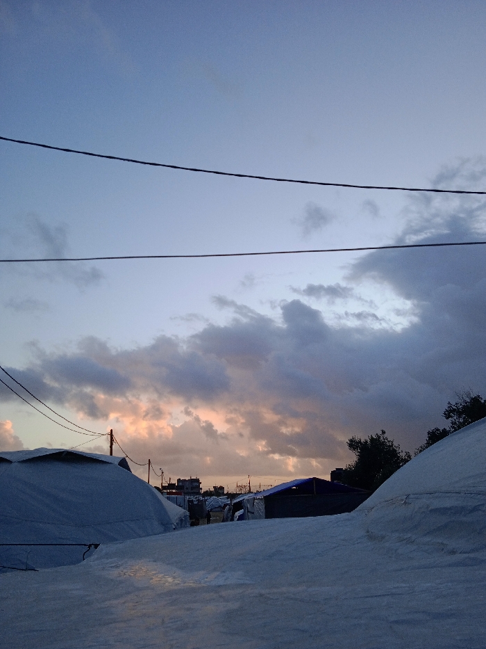 This photograph, taken in Khan Yunis, Palestinian Territory, captures an outdoor scene at either sunrise or sunset, characterized by a dramatic sky and a landscape of temporary shelters. The upper half of the image is dominated by the sky, displaying a blend of soft blue and grey tones, with a prominent band of warm, golden-orange light visible along the horizon, partially hidden by dark, heavy clouds. Several dark utility wires traverse the sky horizontally, suggesting infrastructure.
In the foreground and midground, multiple large, rounded structures, resembling tents or temporary dwellings, are visible. These structures appear to be covered in a light-colored, possibly white or light grey, tarpaulin or similar material, with some surfaces exhibiting a slightly reflective or wet appearance. A significant portion of one such structure occupies the lower left, while another partial one is seen on the lower right. In the background, beyond these primary structures, indistinct buildings or smaller shelters and sparse vegetation are visible. A utility pole stands upright in the midground, further emphasizing human presence and basic services.
No individuals are visible, and there is no discernible text within the image. The atmospheric lighting, contrasting the dark clouds with the warm glow on the horizon, creates a contemplative and somewhat stark mood, highlighting the temporary nature of the depicted settlement under a changing sky.