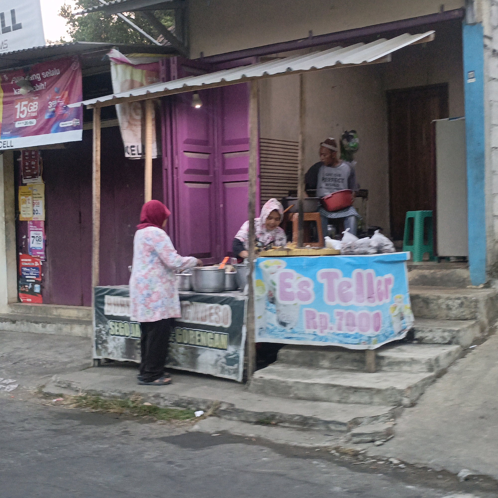 A street-level shot captures a food stall in Salatiga, Indonesia. The stall features a blue banner with pink lettering advertising "Es Teller" for Rp. 7000. Behind the banner, a woman in a pink headscarf and a floral patterned shirt stands at a counter with pots and bowls, attending to her work. To her right, a younger woman with a headscarf, also in a patterned shirt, is seated and appears to be preparing food. A man in the background, wearing a grey t-shirt and a white head covering, is also present. The stall is constructed with a corrugated metal roof and wooden posts, with purple doors visible behind the seated woman. Promotional banners, including one displaying "15 GB," are attached to the structure. The foreground shows a paved walkway and a road, with some grass and debris present.