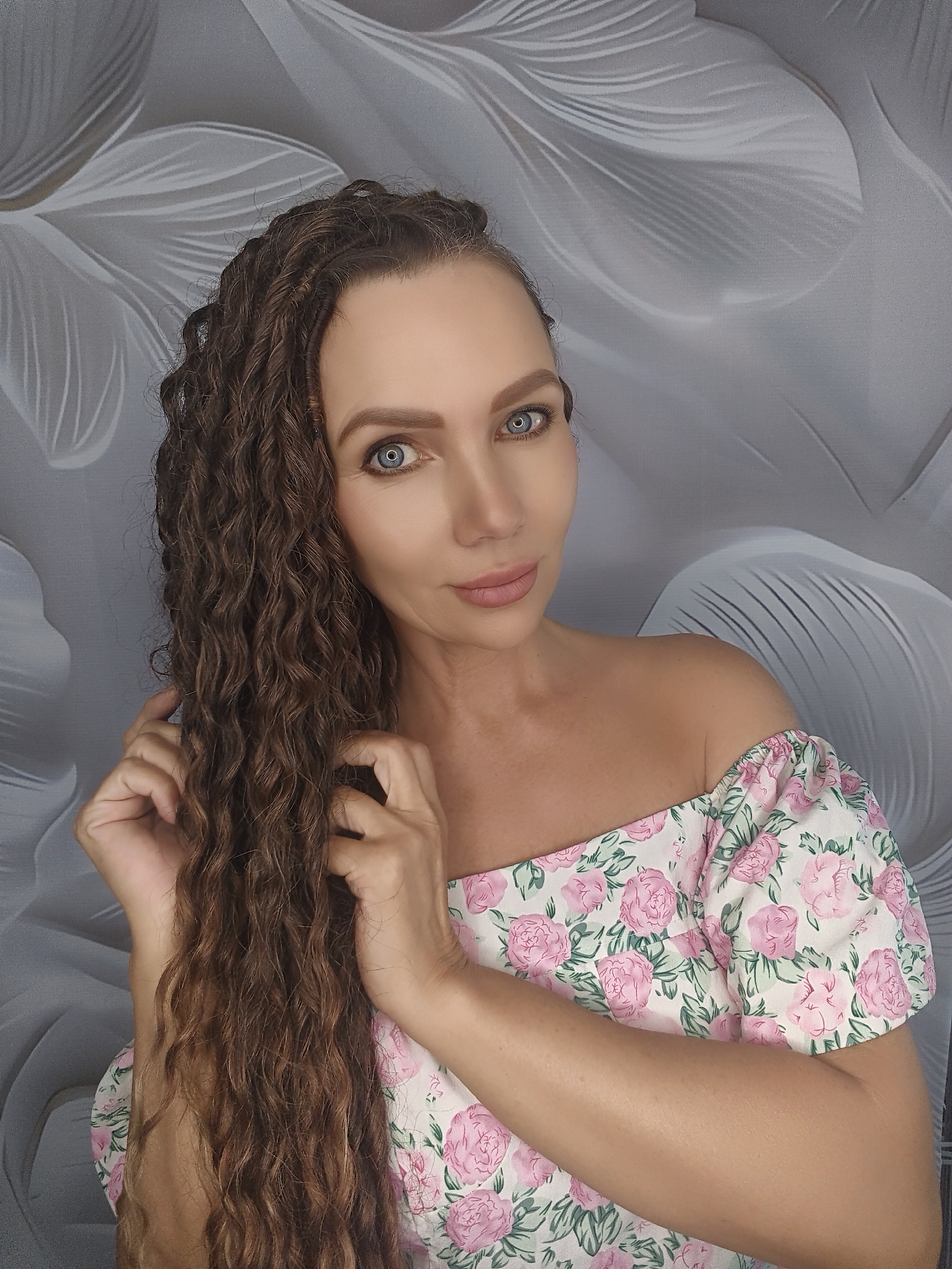 A woman with long, curly brown hair and blue eyes is captured in a portrait-style photo. She is wearing an off-the-shoulder top adorned with a floral pattern of pink roses and green leaves. Her hands are gently holding her hair. The background features a textured gray wallpaper with a subtle, abstract floral design. The lighting is soft, highlighting her features and the details of her clothing and hair.