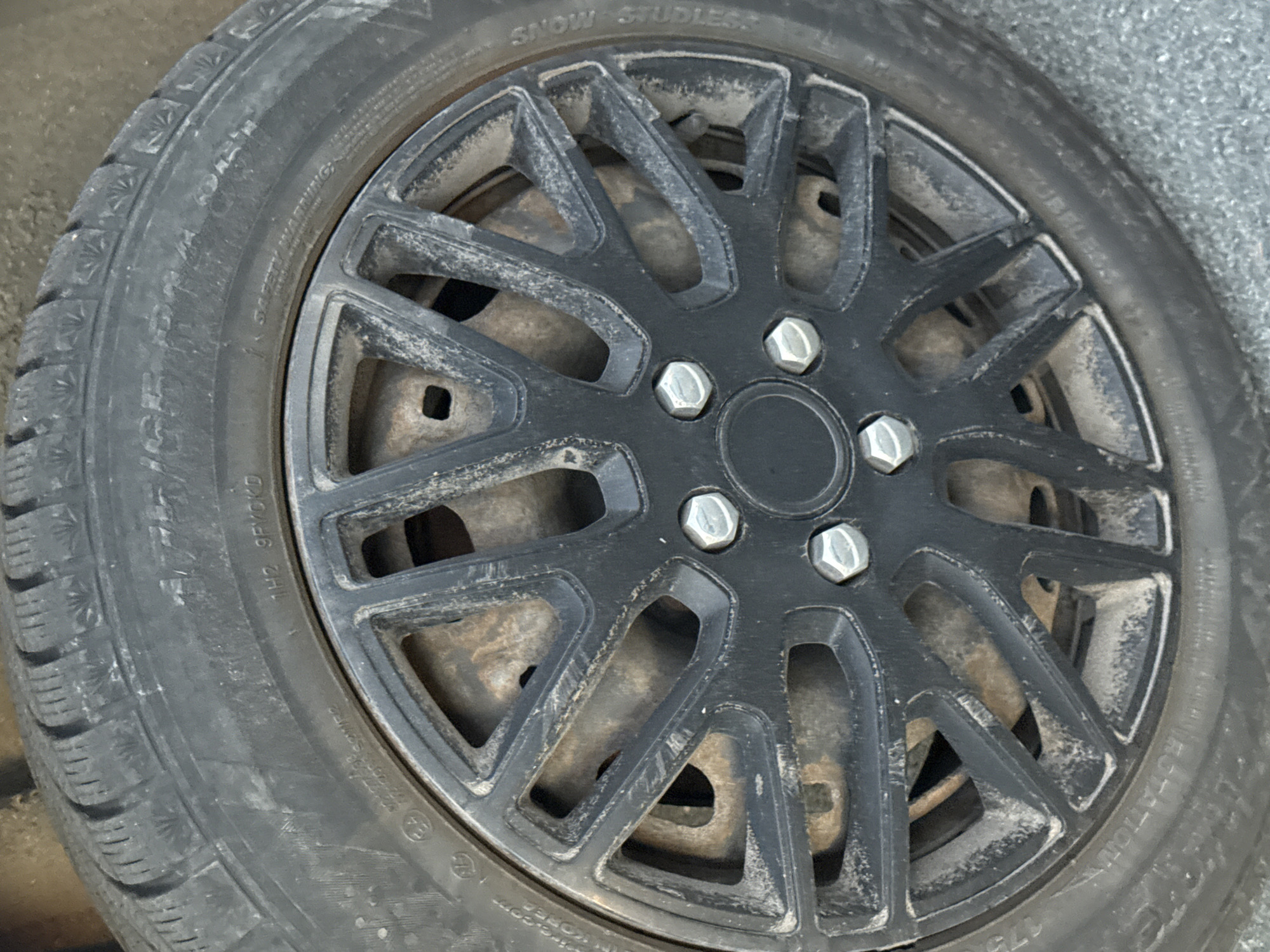 The image shows a close-up, eye-level view of a car's wheel, featuring a black alloy rim with a multi-spoke design. The tire is a dark color and appears to be somewhat dusty. The wheel is mounted on a vehicle parked outdoors on what appears to be an asphalt surface. The scene is devoid of any human presence or activity. The image captures details of the tire's sidewall markings and the lug nuts securing the wheel. There are no specific landmarks or indicators to suggest a location within Tallinn, Estonia.