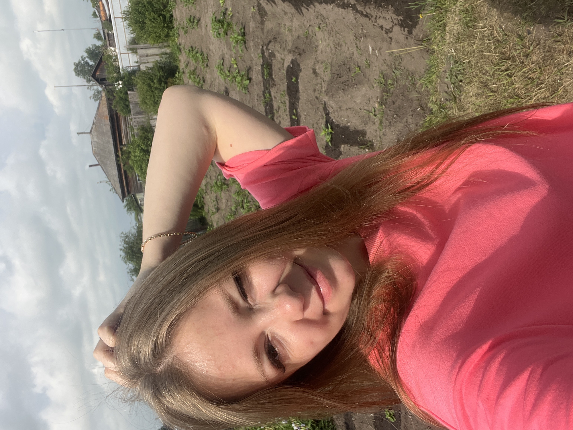 A young woman with long blonde hair is taking a selfie outdoors. She is wearing a pink t-shirt and has her left arm raised, with her hand in her hair. She is winking at the camera, and her right hand is resting on her arm. In the background, there is a garden with rows of plants, a wooden fence, and houses. The sky is cloudy. The location is likely in the village of Sukhrinskoye, Russia, based on the presence of traditional wooden houses and a garden plot, indicative of a rural setting.