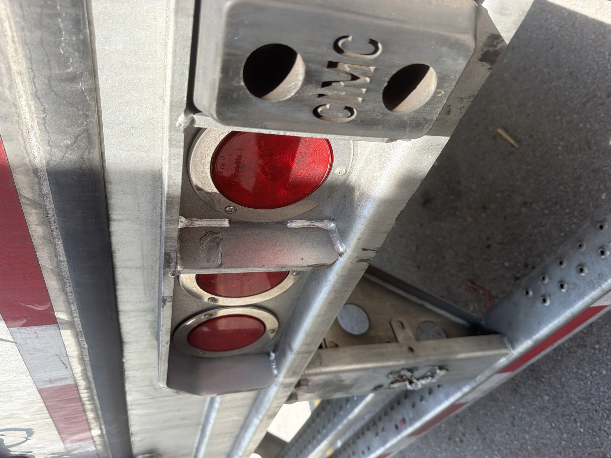 The image shows the rear of a silver trailer, likely a semi-trailer, in what appears to be a commercial or industrial area. The trailer has red and white reflective tape along its lower edge and a black rubber rub rail above the taillight assembly. Three round red LED taillights are visible, recessed into a metal housing. One of the lights has "TOP" and "Grote" branding. To the right of the taillights is a rectangular metal housing with two oval cutouts, one of which displays the embossed letters "CIMC". The ground is asphalt, with some debris visible. The overall impression is a functional, utilitarian vehicle component. The scene's specific location within the city of Lebanon, United States of America, is not discernible from the image alone.