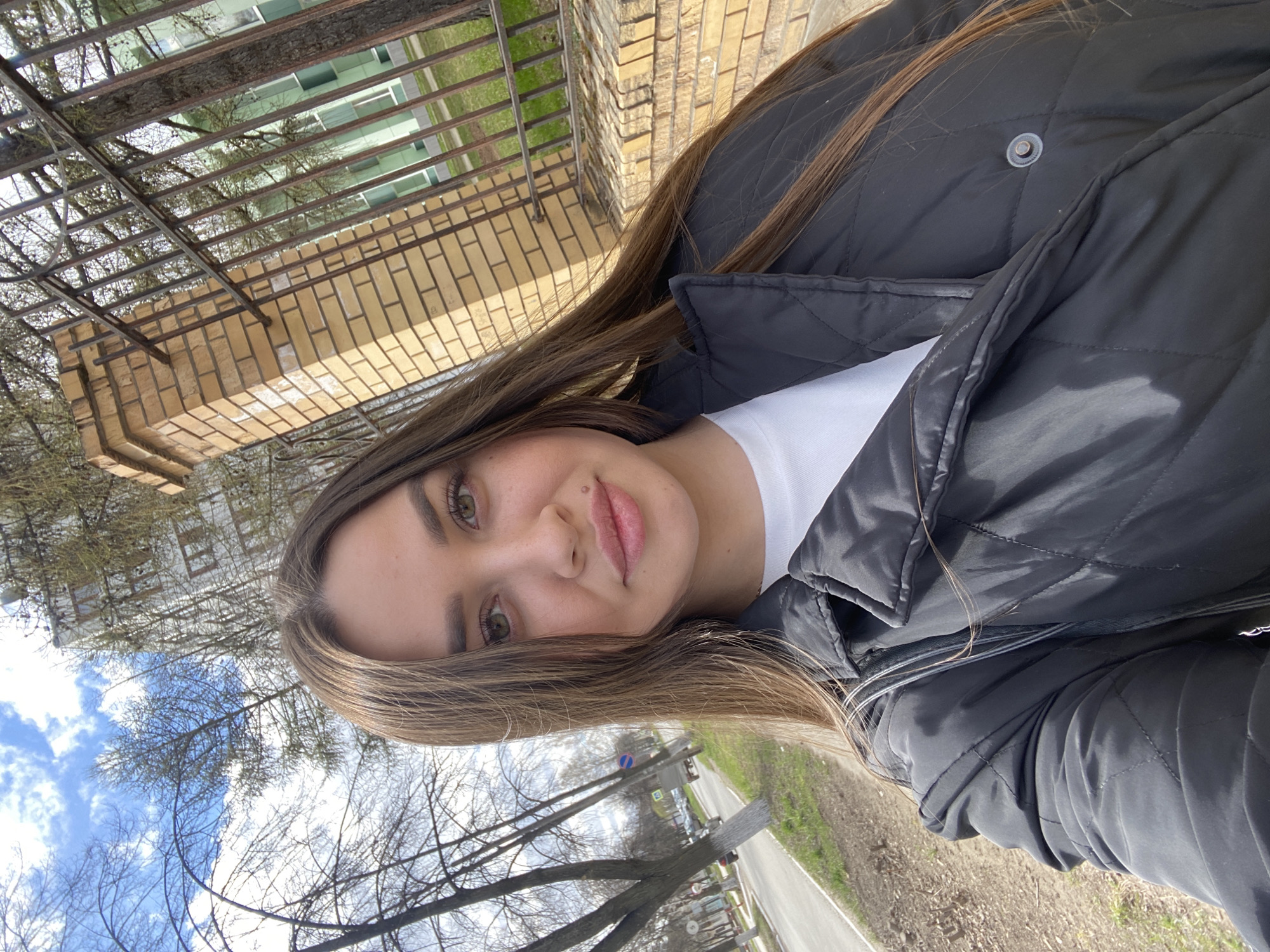 A woman is pictured in an outdoor setting in the city of Chaikovsky, Russia. She wears a black quilted jacket over a white t-shirt. Brown hair falls around her face. Behind her is a brick wall with a metal fence. There are trees and blue sky visible above and beyond the fence. Further behind, there is a street with trees alongside it, along with buildings. The photograph appears to have been taken at ground level.