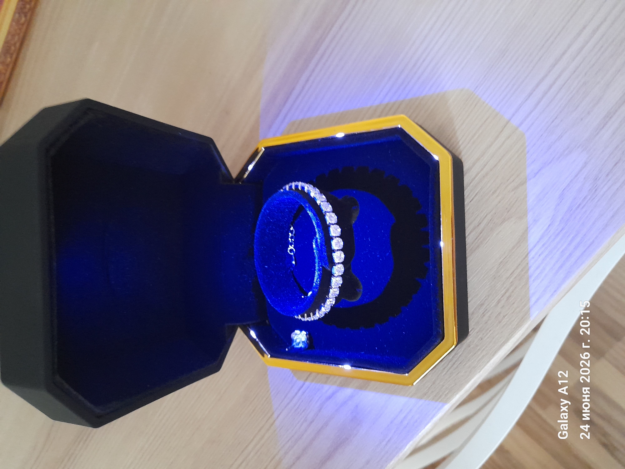 The image shows an opened jewelry box, which is black and hexagonal with a gold trim. Inside, the box is lined with dark blue velvet and contains a sparkling diamond bracelet arranged in a circular fashion on a velvet stand. To the left of the bracelet, a small, single diamond is visible. The jewelry box is placed on a light-colored wooden surface. The lower left corner of the image displays a timestamp and device information: "Galaxy A12" and "24 июня 2026 г. 20:15". The context of Kara-Köl City, Kyrgyzstan is not depicted in the image.
