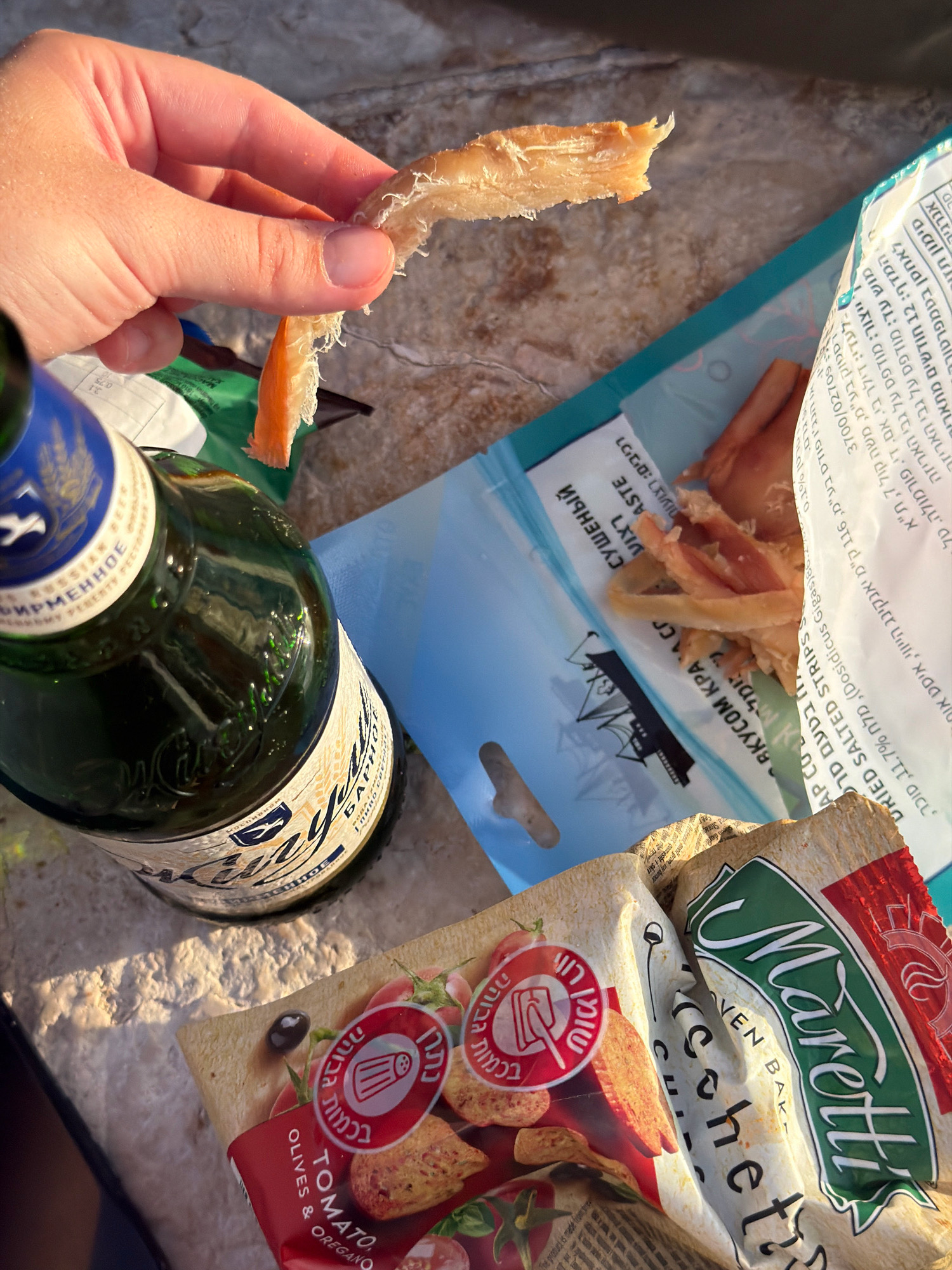 In Tel Aviv, Israel, a hand holds a piece of dried fish. Beside it, a bottle of Russian beer, a bag of dried squid strips, and a bag of Maretti bruschette with tomato and oregano flavor are scattered on a textured surface that appears to be a light-colored stone. The scene is lit by natural sunlight, casting shadows and highlighting the textures of the food items and the surface.